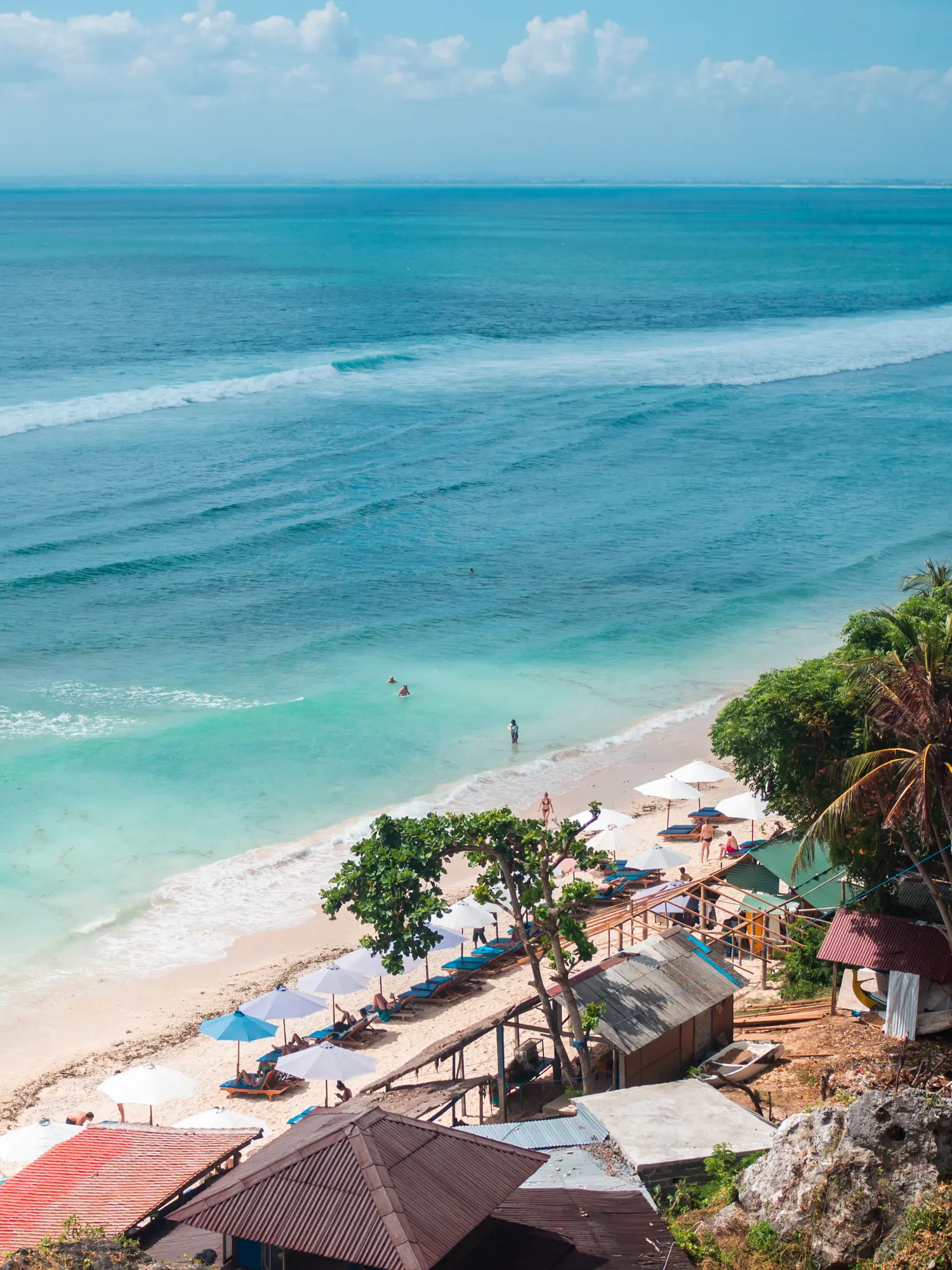 View from the cliff above of the white Thomas Beach and blue water lined by jungle and palm trees, one of the best beaches in Uluwatu Bali.