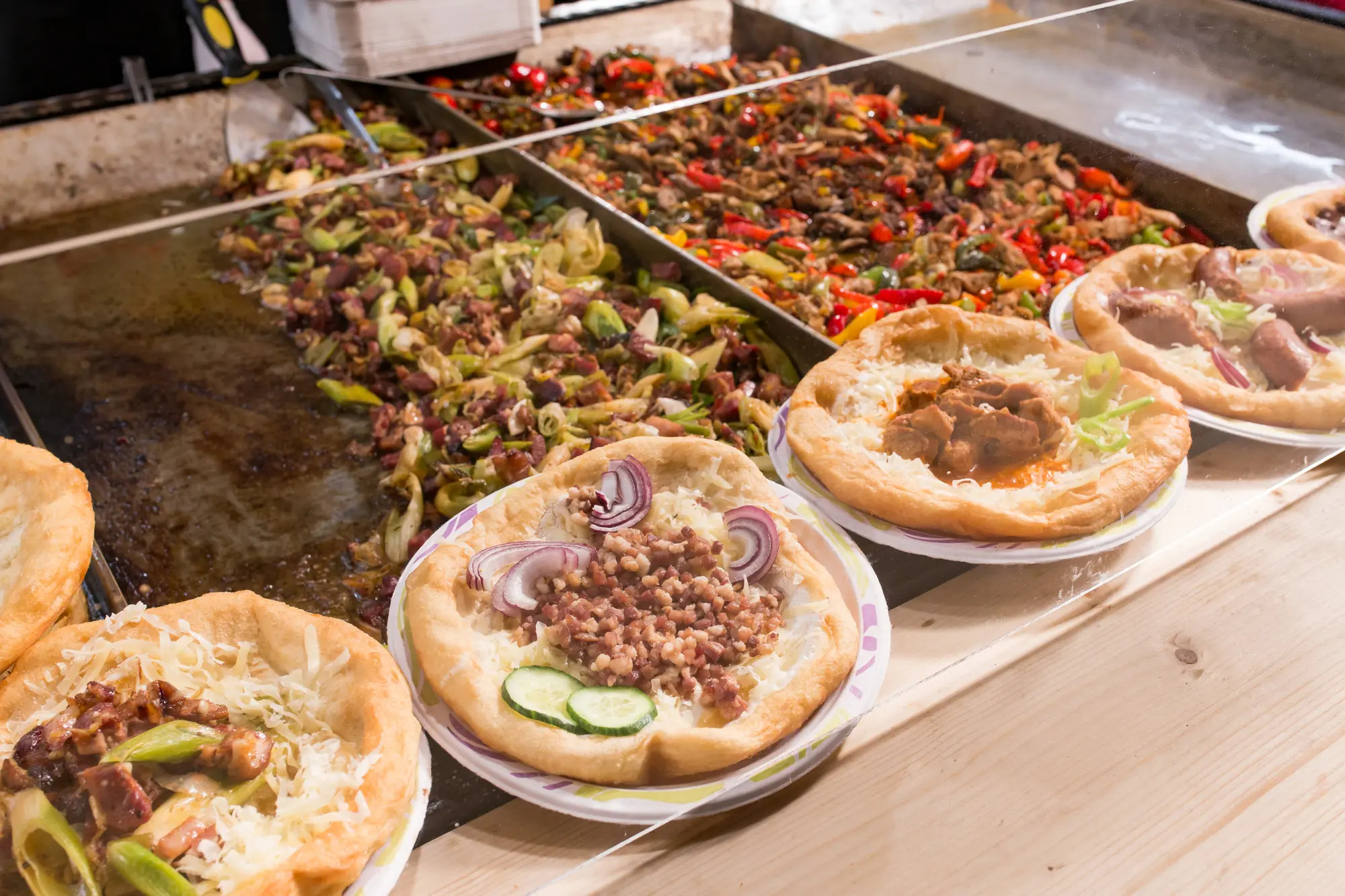 Lángos with different toppings in front of two grills with meat and vegetables at a street food stall in Budapest. 