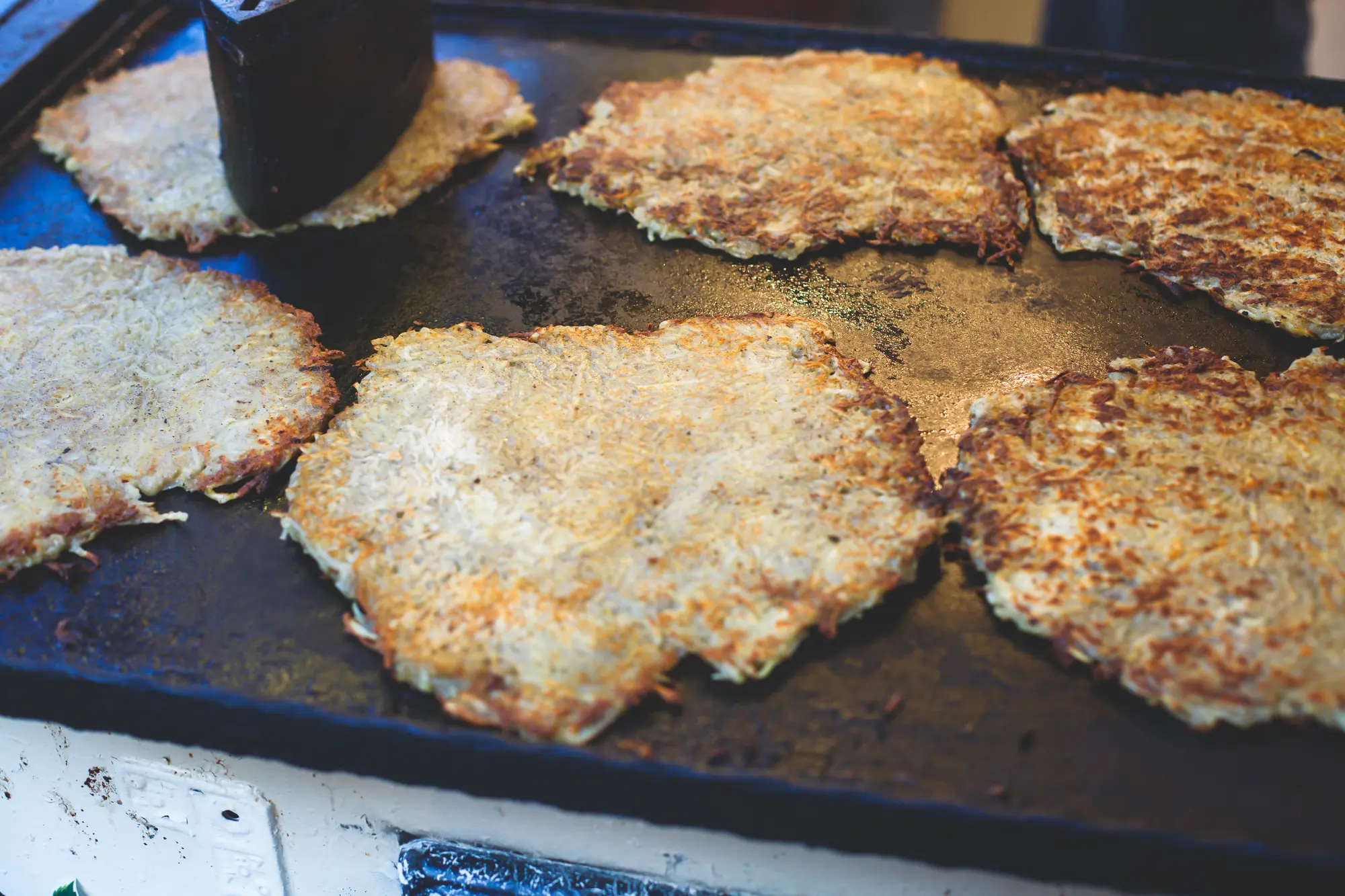 Six large Latke (potato pancakes) on a black grill at a street food market in Budapest.