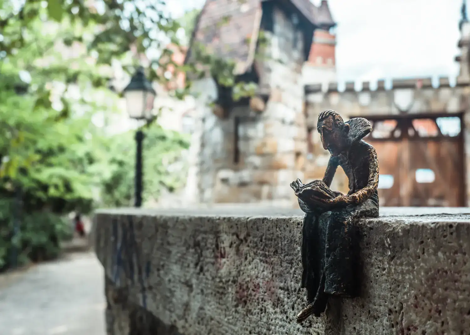 Mini statue of Dracula sitting on a stone railing while reading a book with a parts of a castle in the background, made by artist Kolodko, a hidden gem in Budapest.