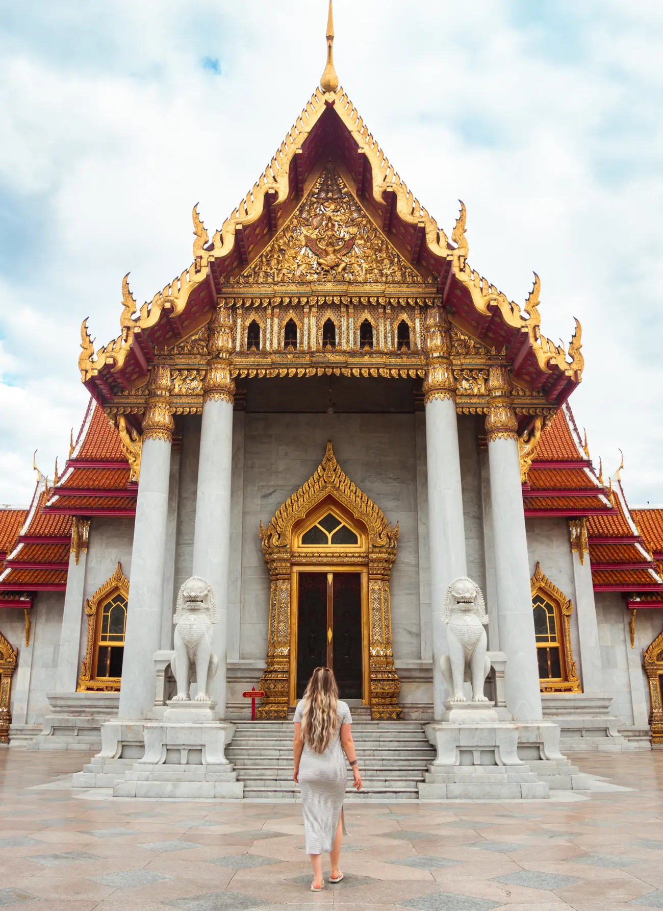 Girl with long hair wearing a maxi dress walking towards Wat Ben Marble temple with red roof and gold details in Bangkok - Indonesia vs Thailand.