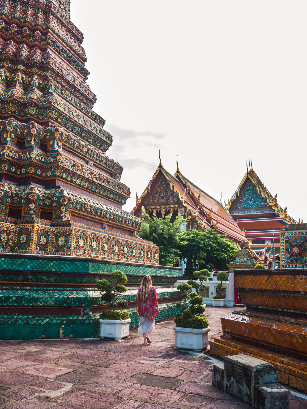 Girl wearing a red kimono walking between two potted trees inside the colorful Wat Pho in Bangkok early in the morning - Indonesia vs. Thailand.