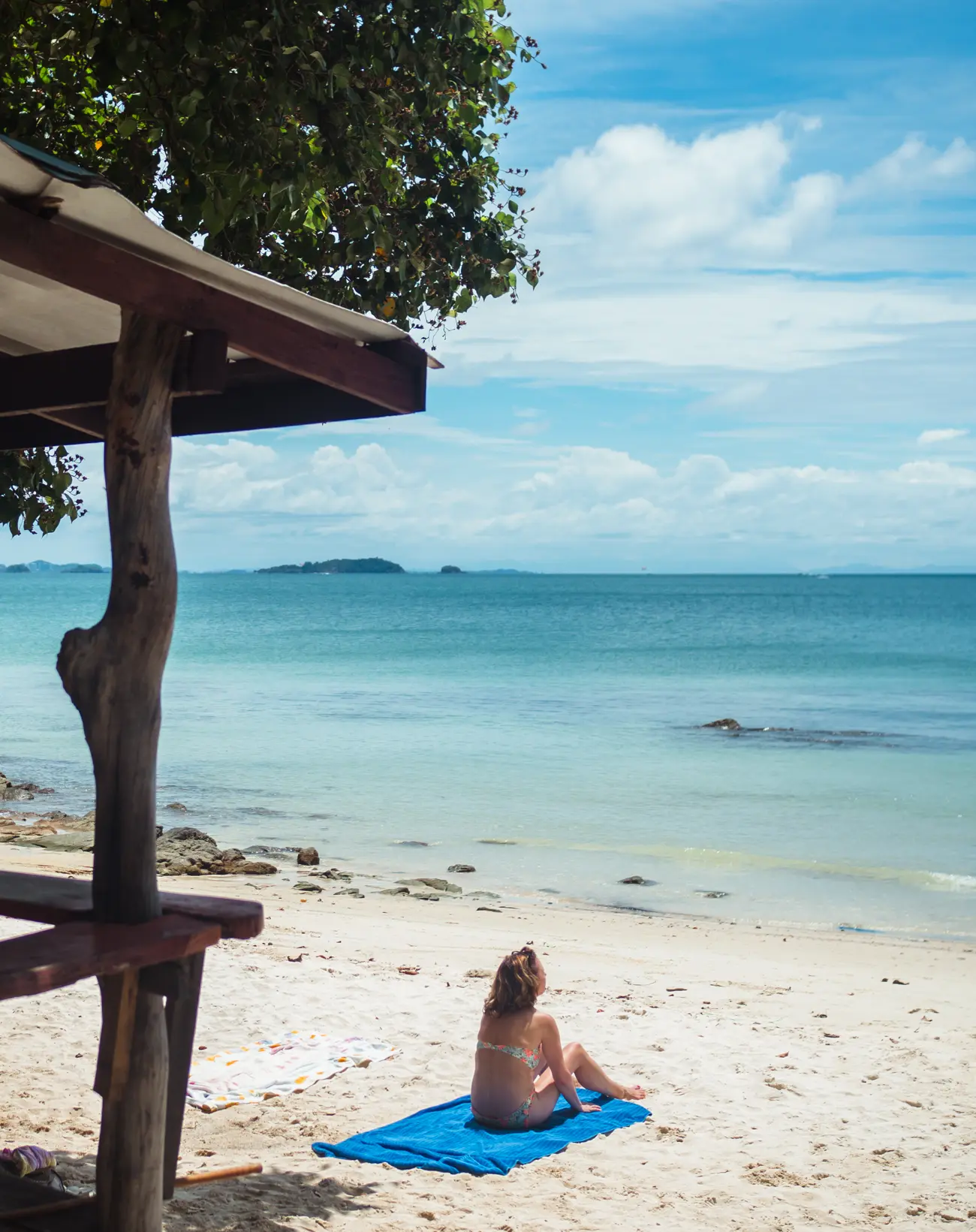 Woman sitting on a blue towel in front of a wooden beach bungalow looking out at the turquoise water on Koh Samet - Indonesia vs. Thailand beaches