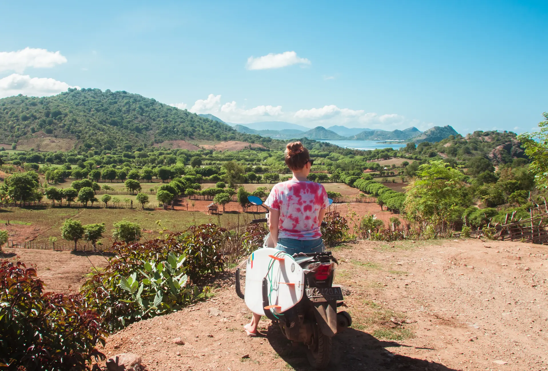 Girl on a scooter with a white surfboard looking out over lush nature towards the ocean in Sumbawa - Indonesia vs. Thailand transportation