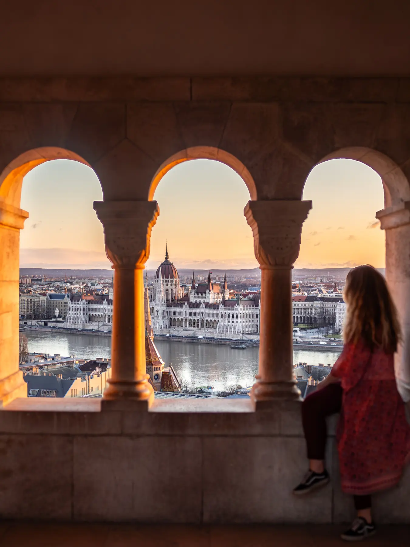 Girl in a pink kimono sitting in one og three arches at Fisherman's Bastian looking out over Budapest Parliament and hidden gems during sunrise.