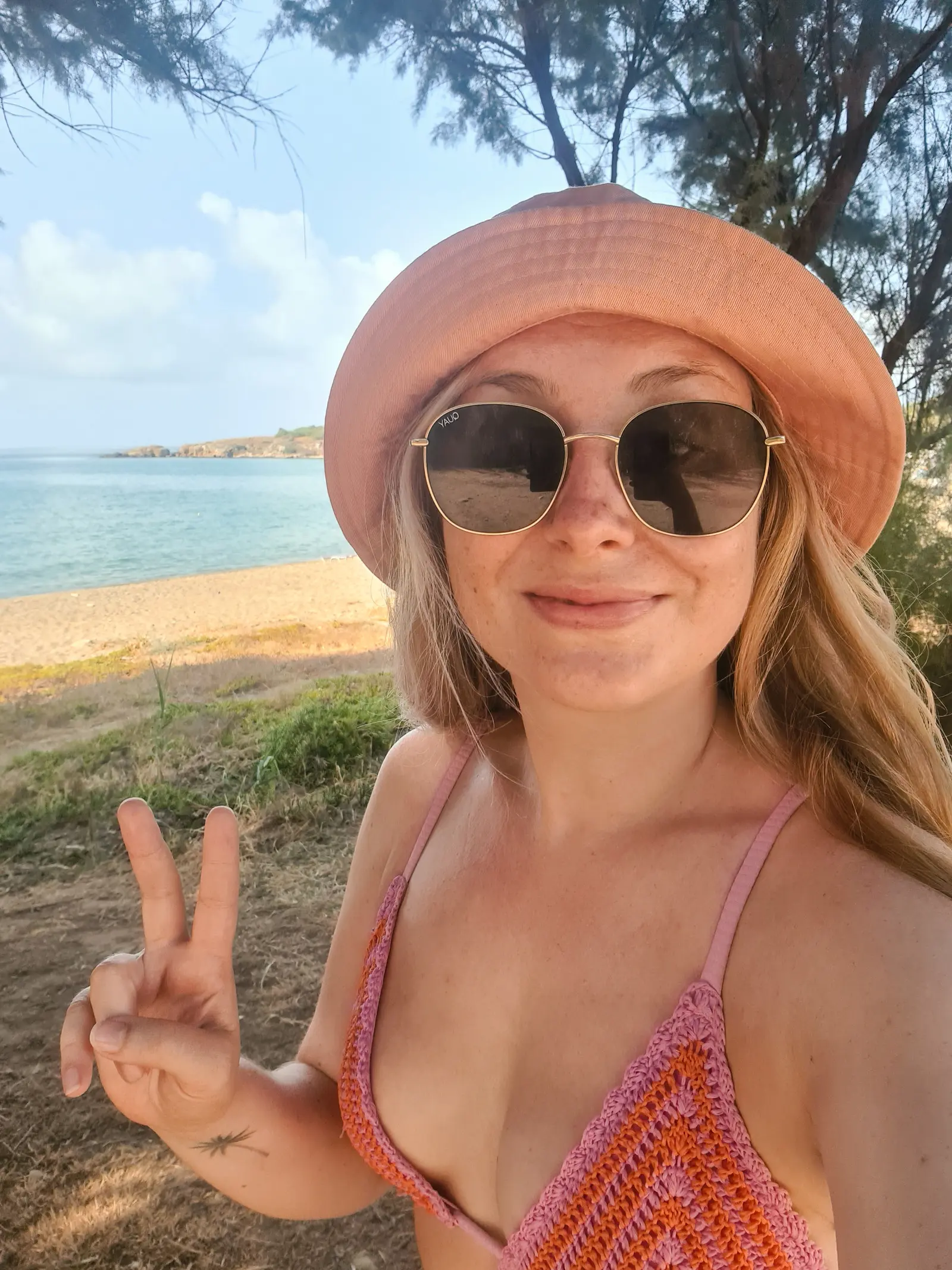 Girl in a orange bucket hat, large round sunglasses and purple bikini top making a peace sign in front of Chrissi Akti Beach, one of the best beaches in Chania Crete.