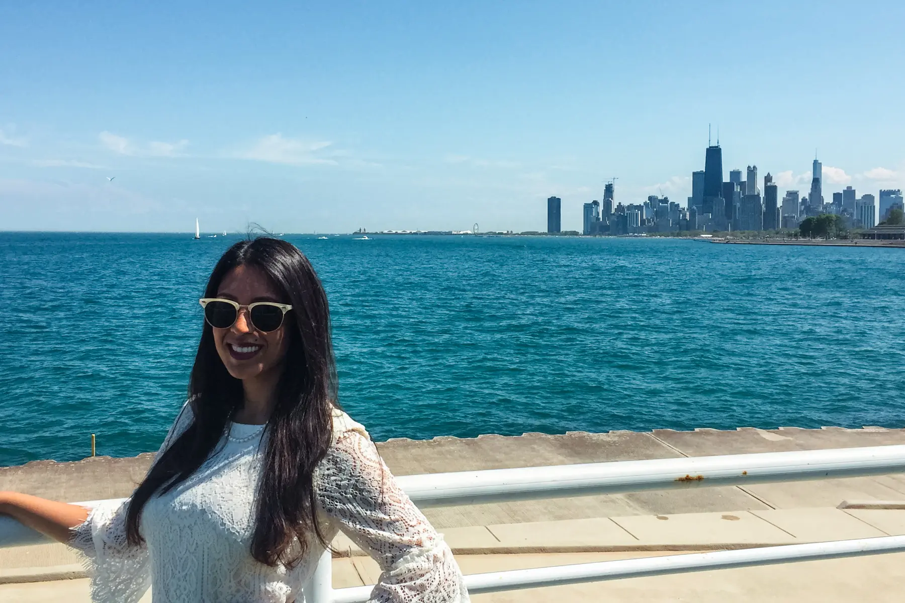 Woman with black hair, wearing a white top and sunglasses, posing on the Lakefront Trail with the skyline in the background, one of the best things to do on a weekend in Chicago.