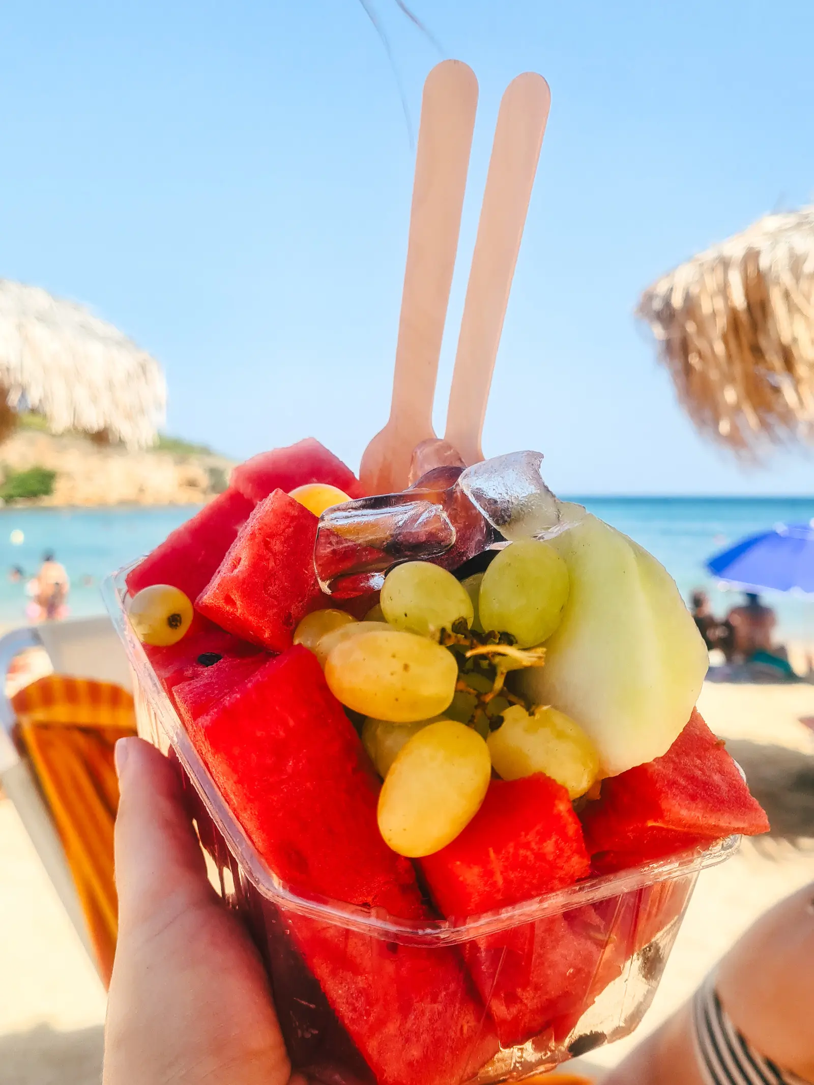 Hand holding a a plastic container with watermelon, grapes, apples, ice cubes and two wooden forks in front of Agii Apostoloi Beach in Chania.