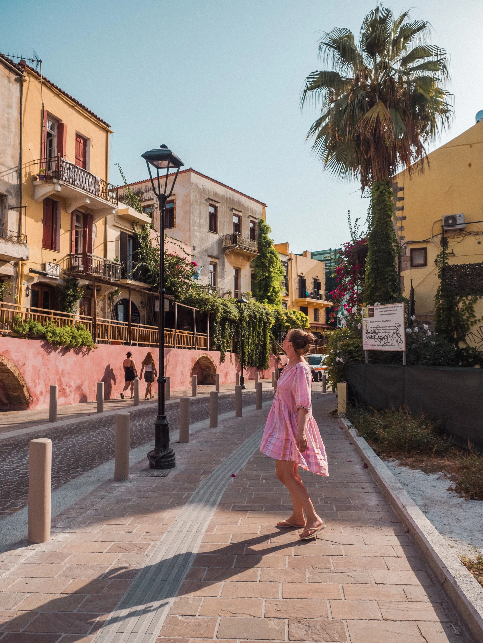 Girl in a pink dress walking up the streets of Chania old town with a palm tree and yellow buildings in the background, one of the top things to do in Chania.