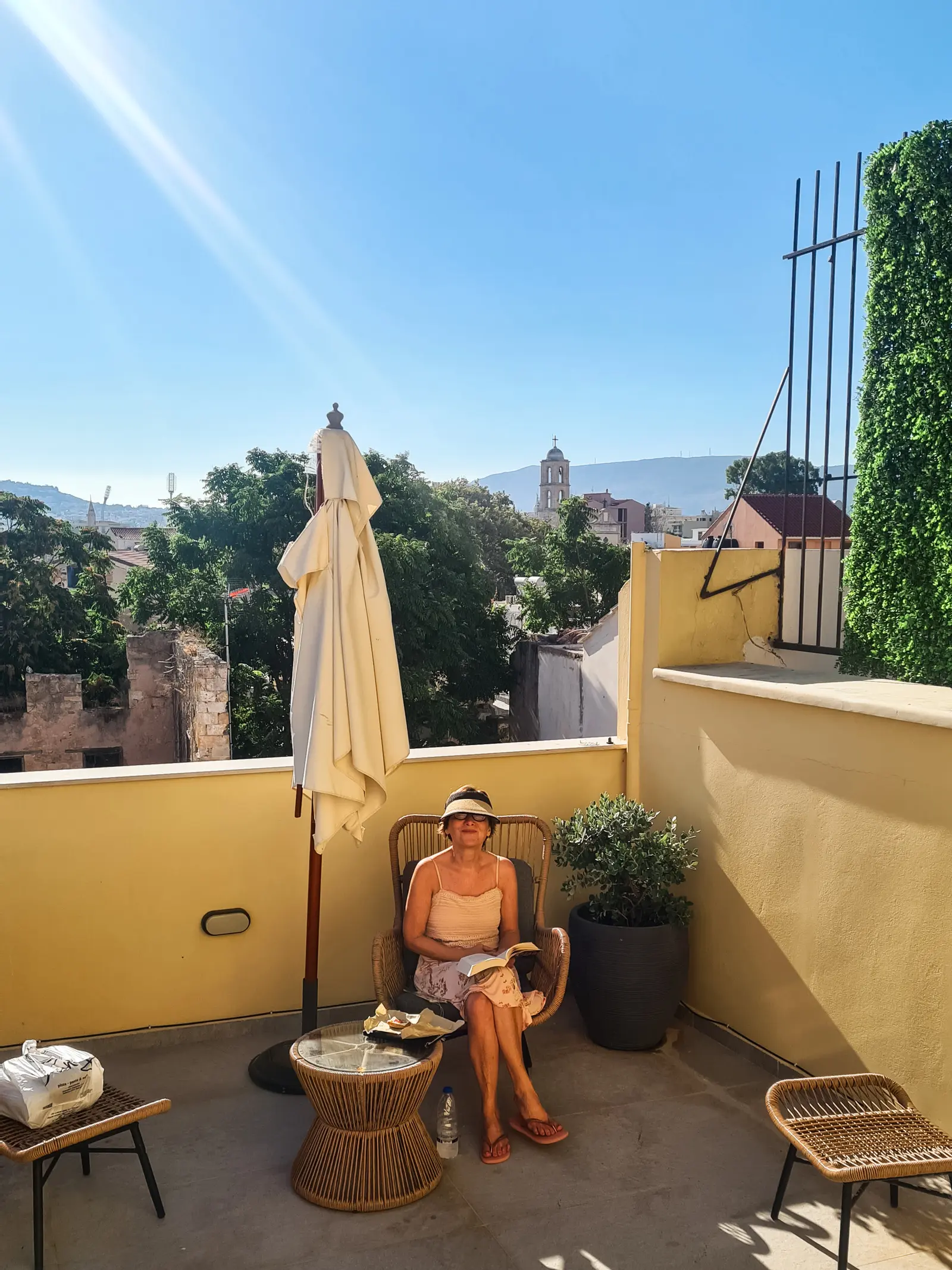 Woman wearing a visor sitting reading a book on a yellow balcony on a sunny morning, one of the best places to stay in Chania.