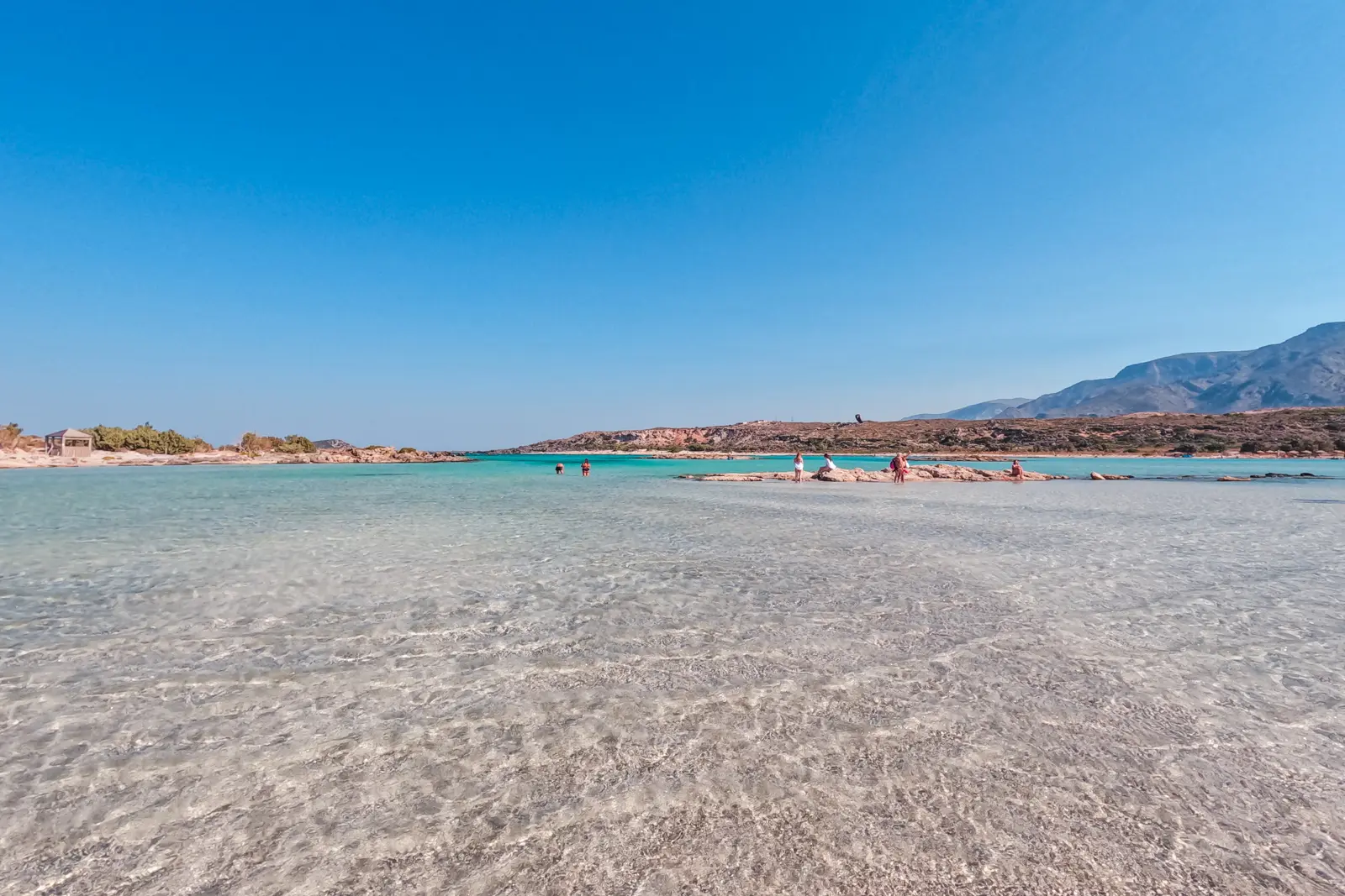 People walking far away in the vast shallow lagoon of Elafonisi Beach on a sunny day with a mountain in the background. Chania to Elafonisi Beach.