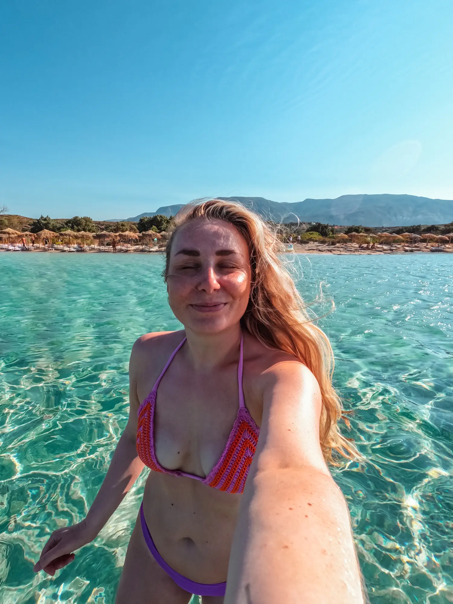 Girl with long hair in a purple and orange bikini taking a selfie with eyes closed in the turquoise waters of Elafonisi Beach from Chania.