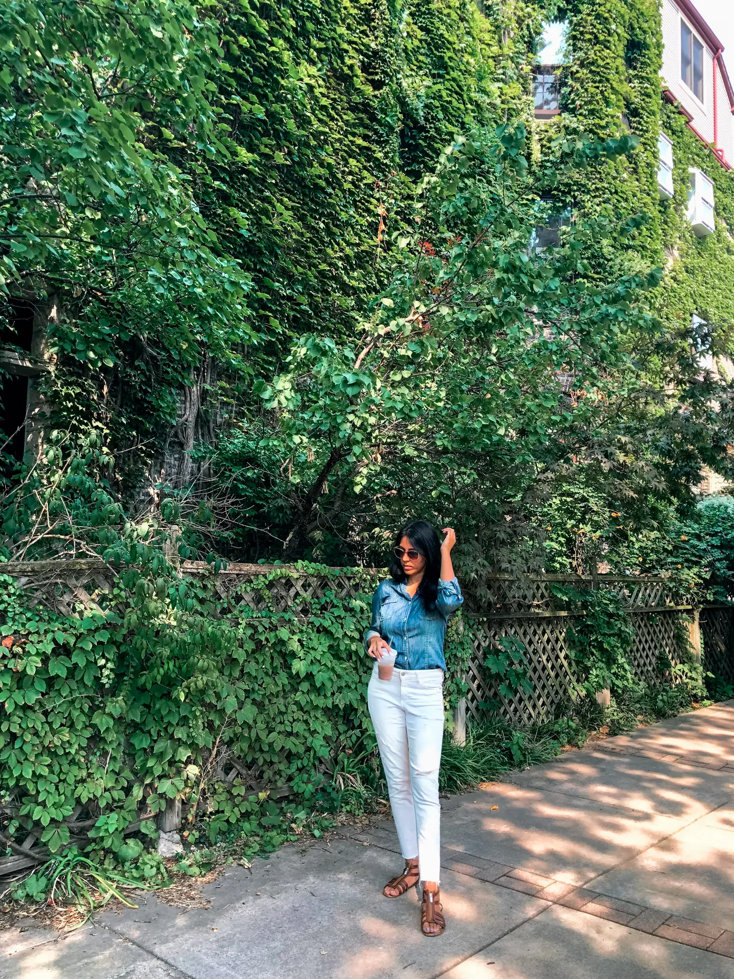 Woman with black hair, wearing white pants and a denim shirt, standing in front of a wall covered in greenery after brunch in Lakeview, one of the best things to do on a weekend in Chicago.