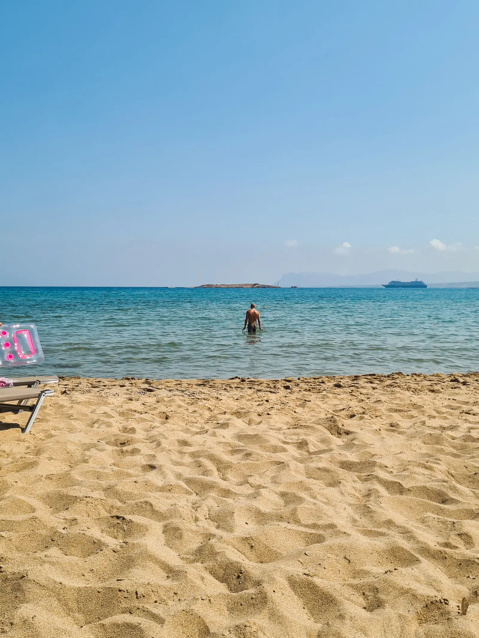 Man standing in shallow blue water in front of the golden sand at Chrissi Akti Beach, one of the closest beaches to Chania.