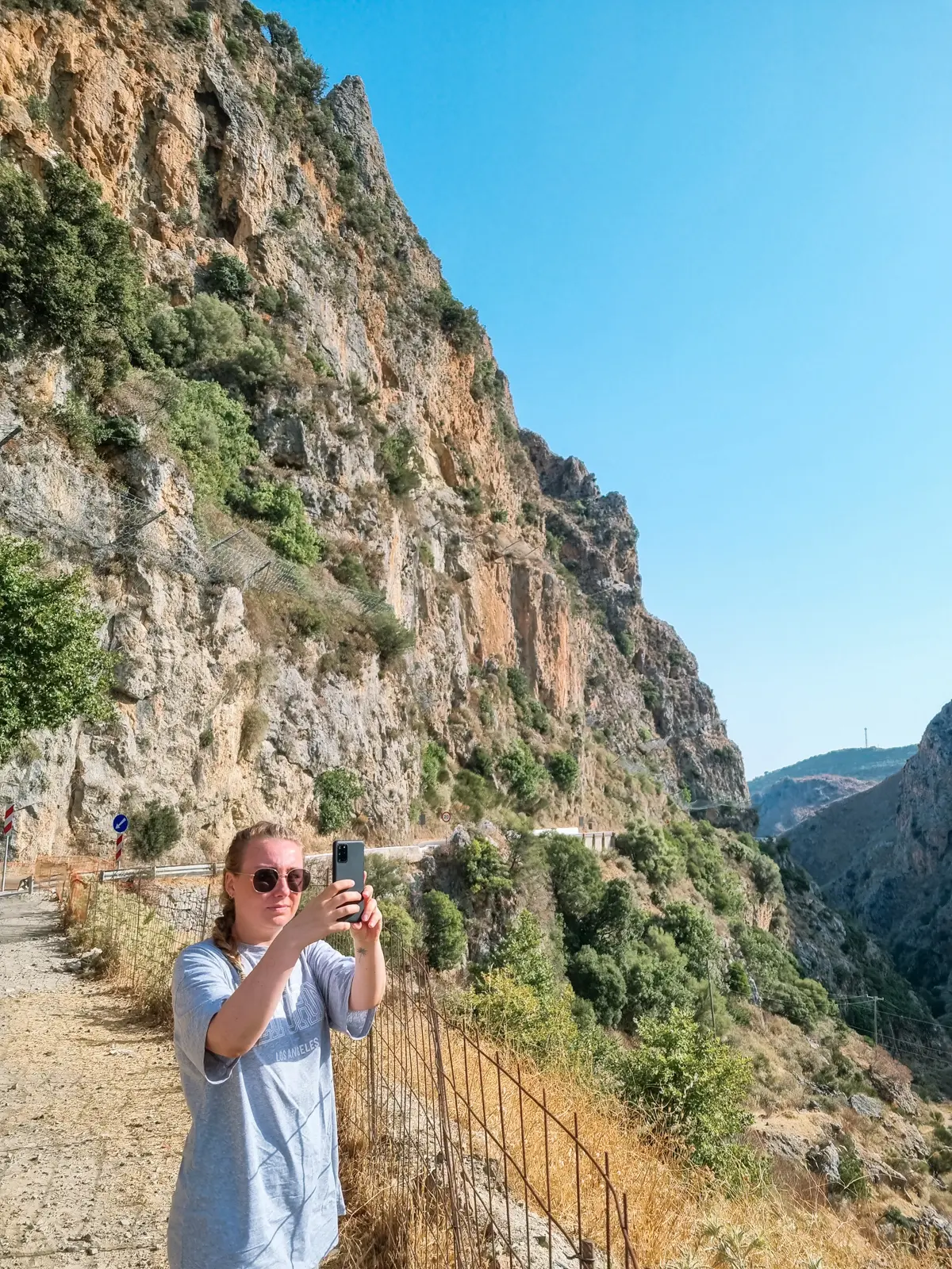 Girl in a grey t-shirt taking photos in front of a mountain with green bushed on the road from Chania and Falasarna to Elafonisi Beach.
