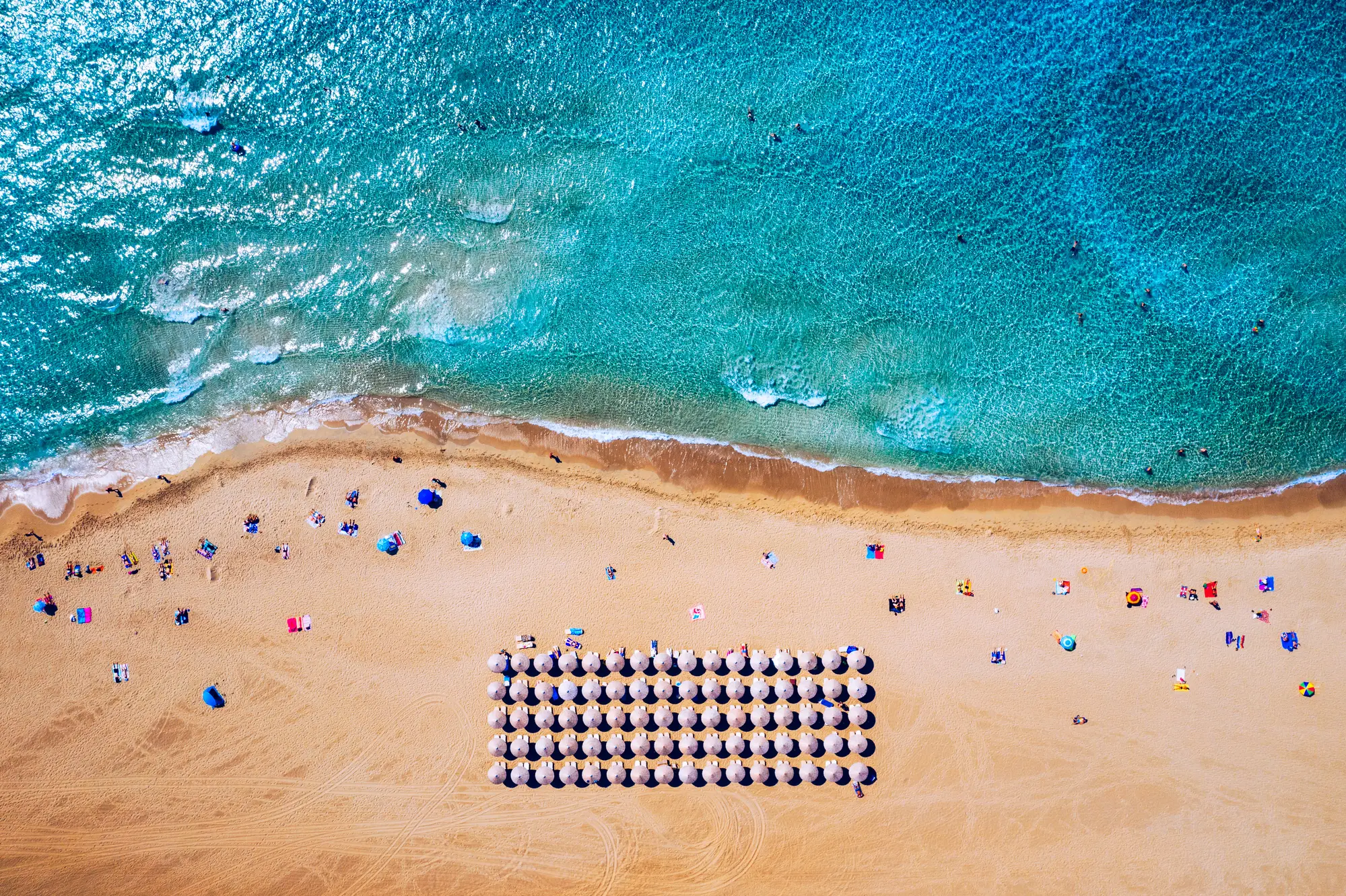 Drone view of a rectangle section of sunbeds and parasols on the golden Falassarna Beach lined by turquoise water in Crete.