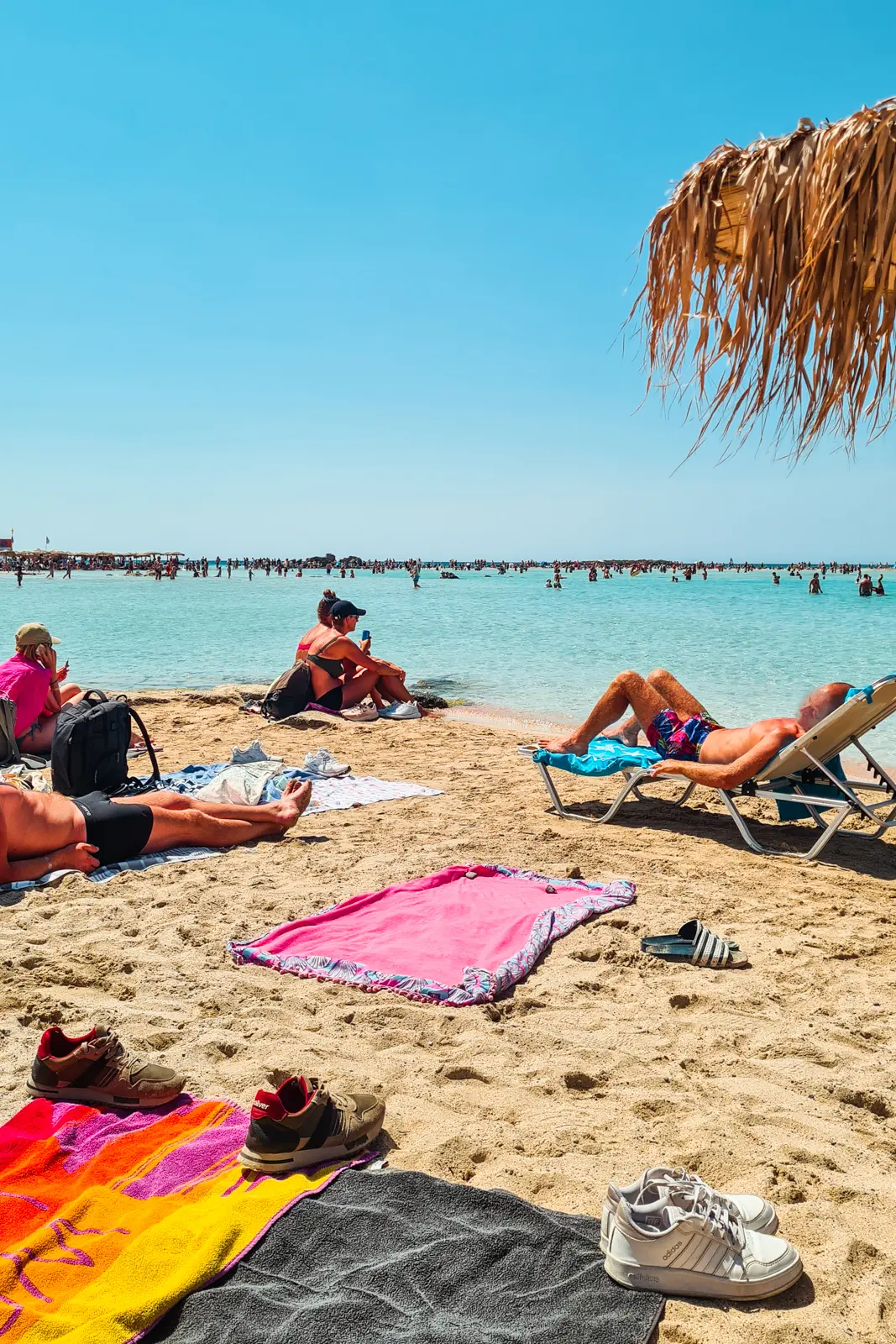 People sitting on towels and sun loungers looking out over the shallow water at Elafonisi Beach on a sunny day in August.