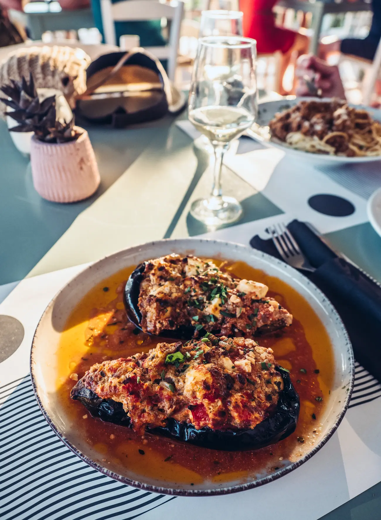 White plate with two half gratinated eggplants with a glass of white wine in the background at Elafonisi Resort by Kalomirakis Family Restaurants at Elafonisi Beach in Crete.