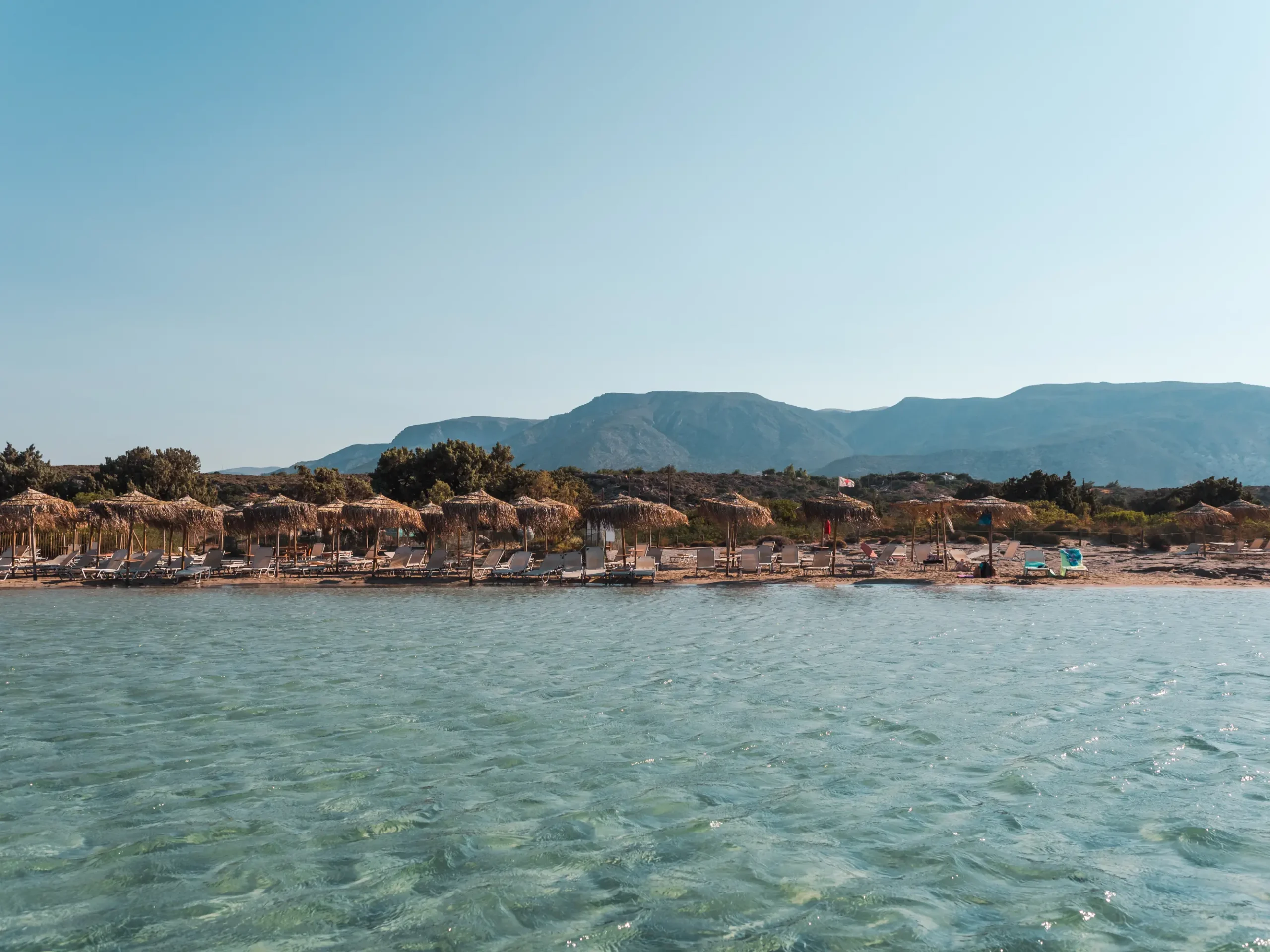 A row of white sun loungers and straw parasols and shallow ocean on a sunny morning at Elafonissi Beach in Crete.