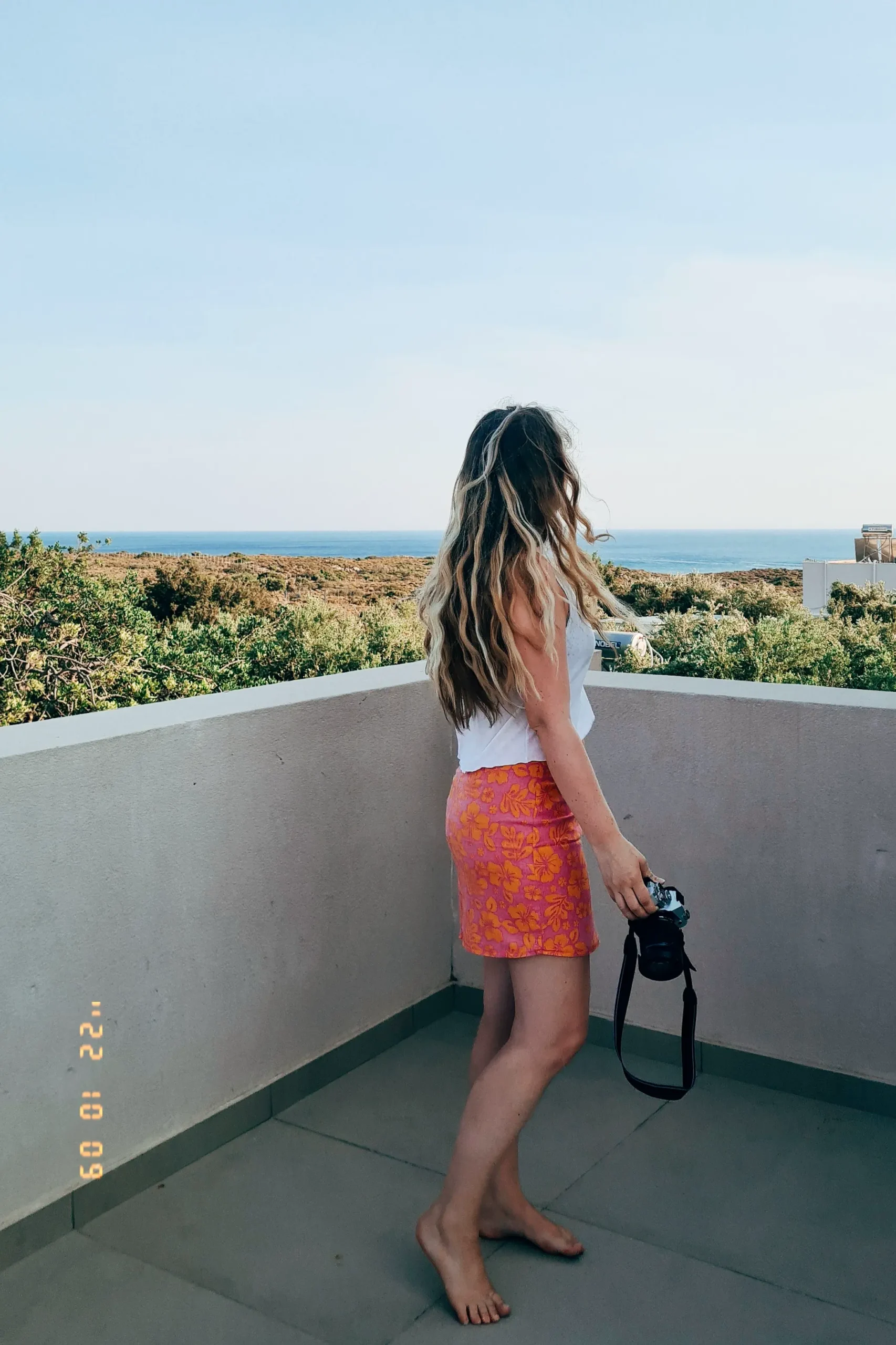 Girl in pink skirt and white top, with long dark blonde hair, holding a camera on the balcony of Elafonisi Resort by Kalomirakis Family.