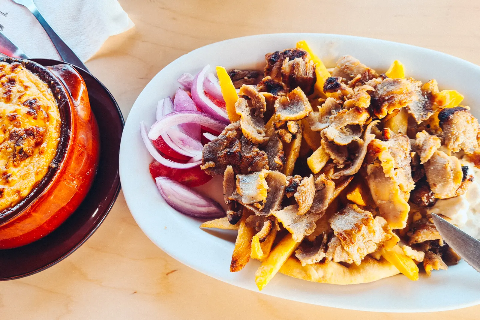 White plate with fries, red onion, tomato and gyros meat. To the side a red ramiken with Moussaka at Panorama Restaurant close to Elafonisi Beach.