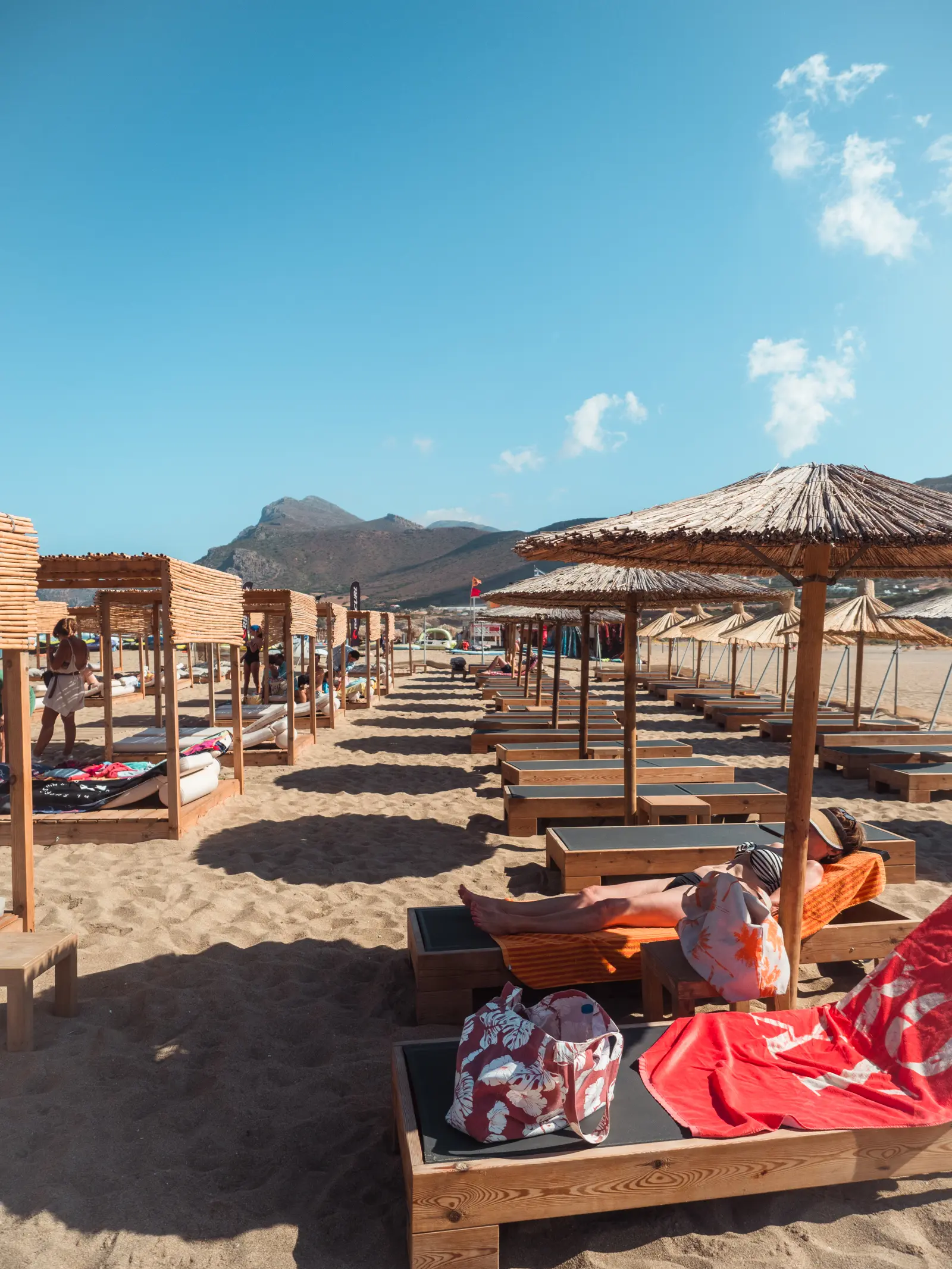 Row of sunbeds with straw parasols to the right and row of wooden cabanas to the left on Falassarna Beach in Crete.