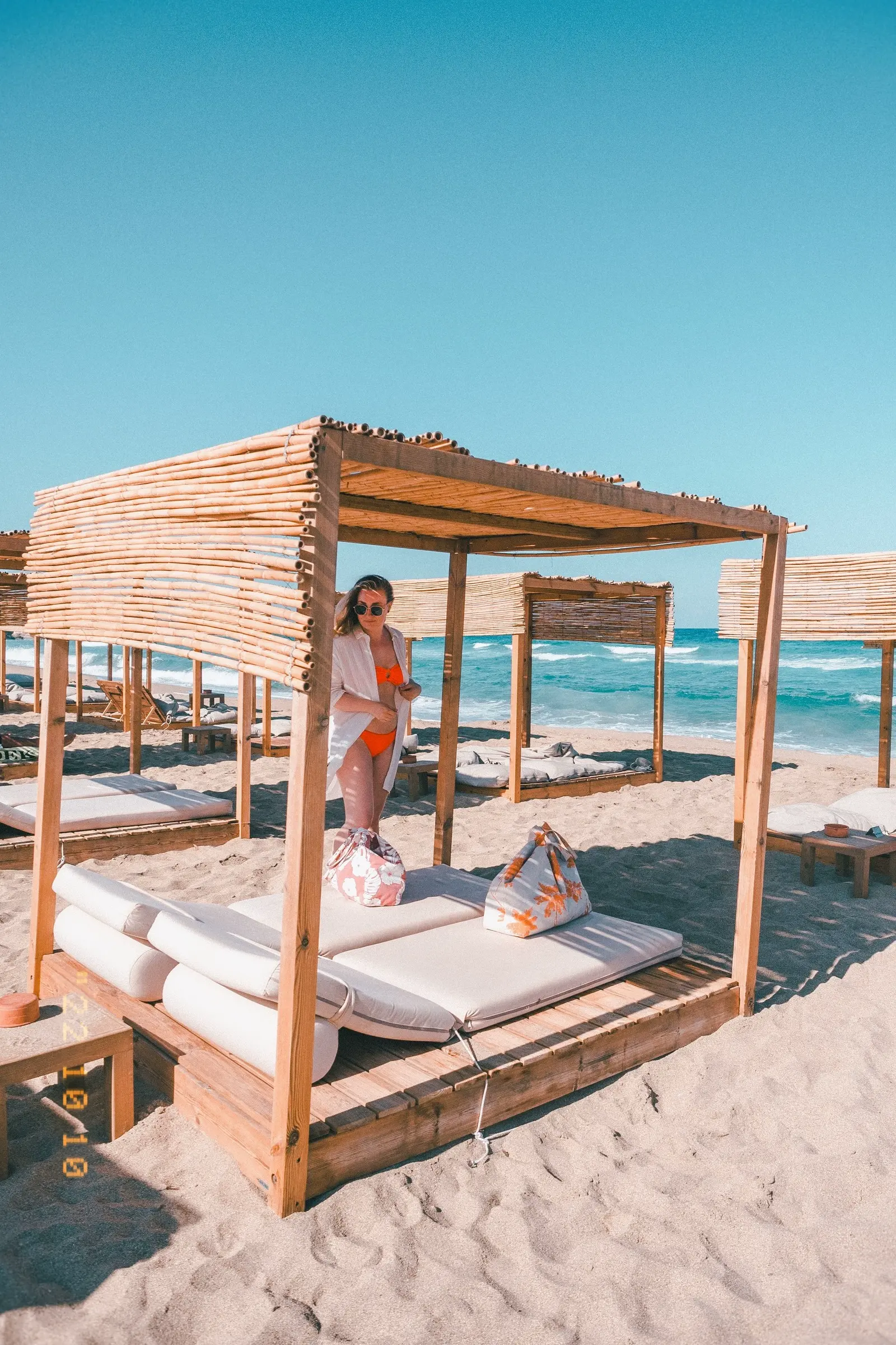 Girl in a white shirt and orange bikini standing behind a wooden cabana on Falassarna Beach in Crete.