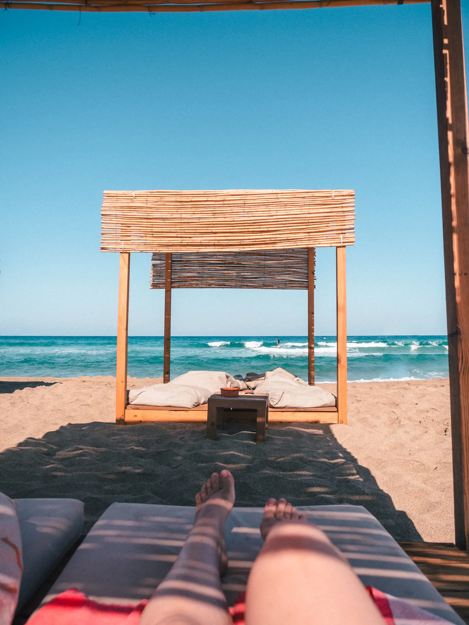 Legs on a red towel inside a cabana looking towards a wooden cabana with a surfer on a wave in the background and Falassarna Beach in Crete.