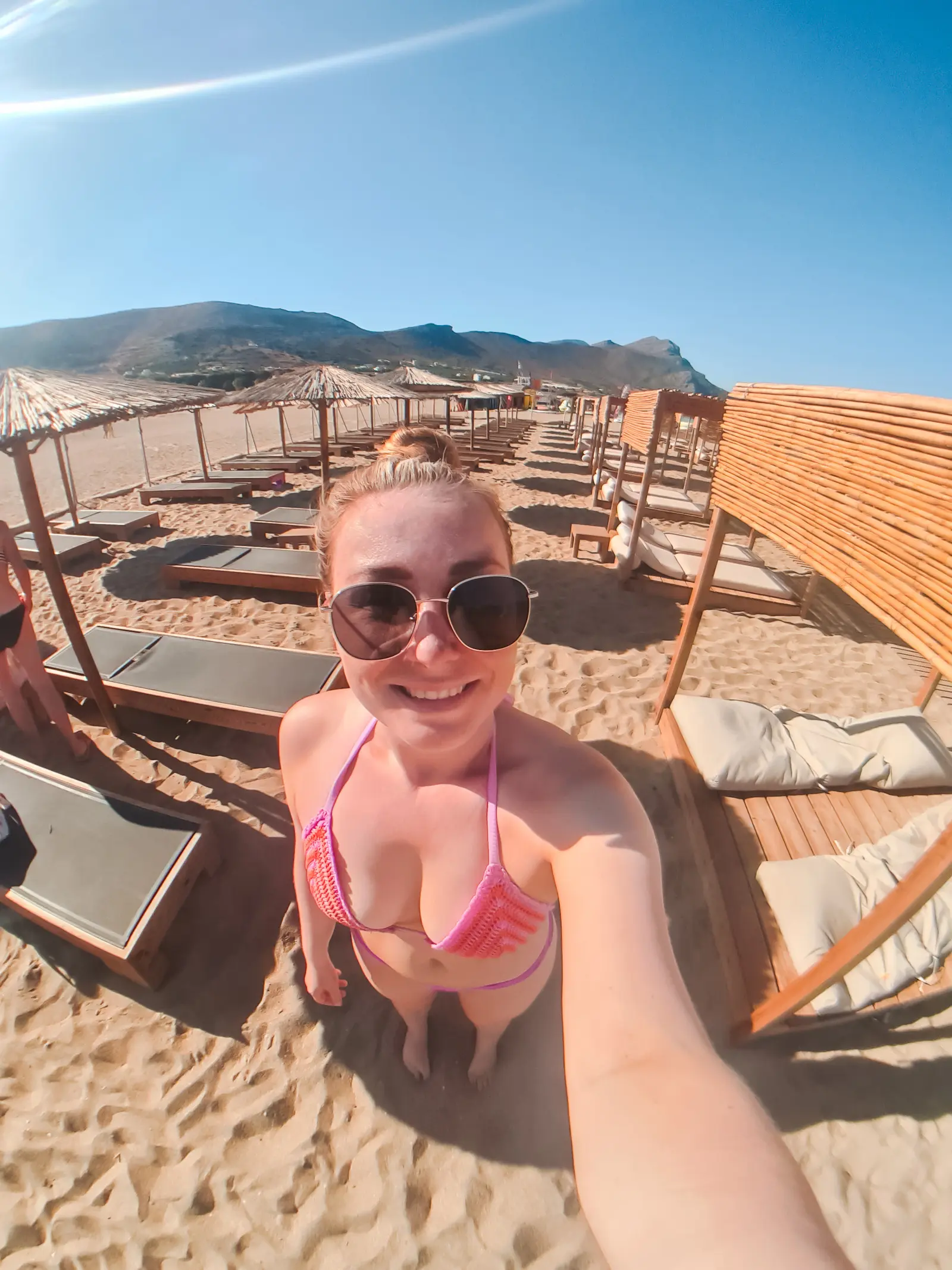Girl with large circular sunglasses, wearing a purple bikini, smiling while taking a selfie with cabanas and sunbeds at Falassarna Beach in the background.
