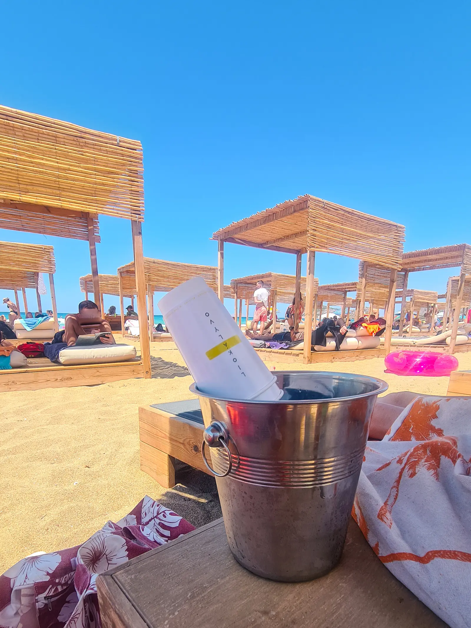 Metal champagne bucket with water and paper glasses standing on a table next to sunbeds and wooden cabanas on Falassarna Beach in Crete.