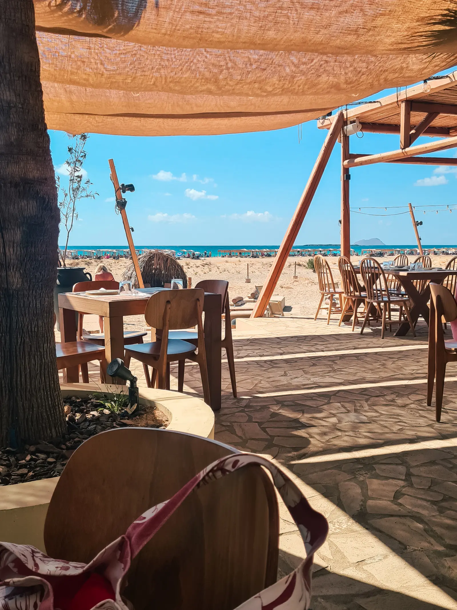 Wooden tables on a stone floor under a cloth roof looking out over Falassarna Beach and the ocean.