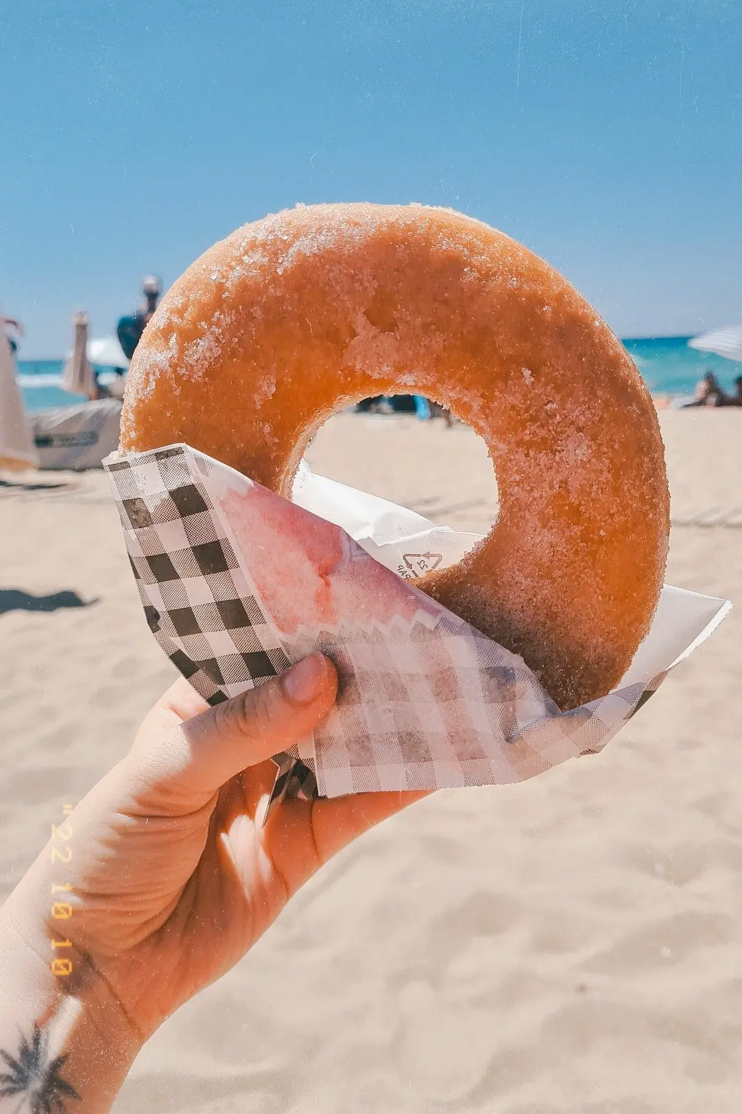 Close up of hand holding a large donut wrapped in white and black checkered paper on Falassarna Beach in Create.