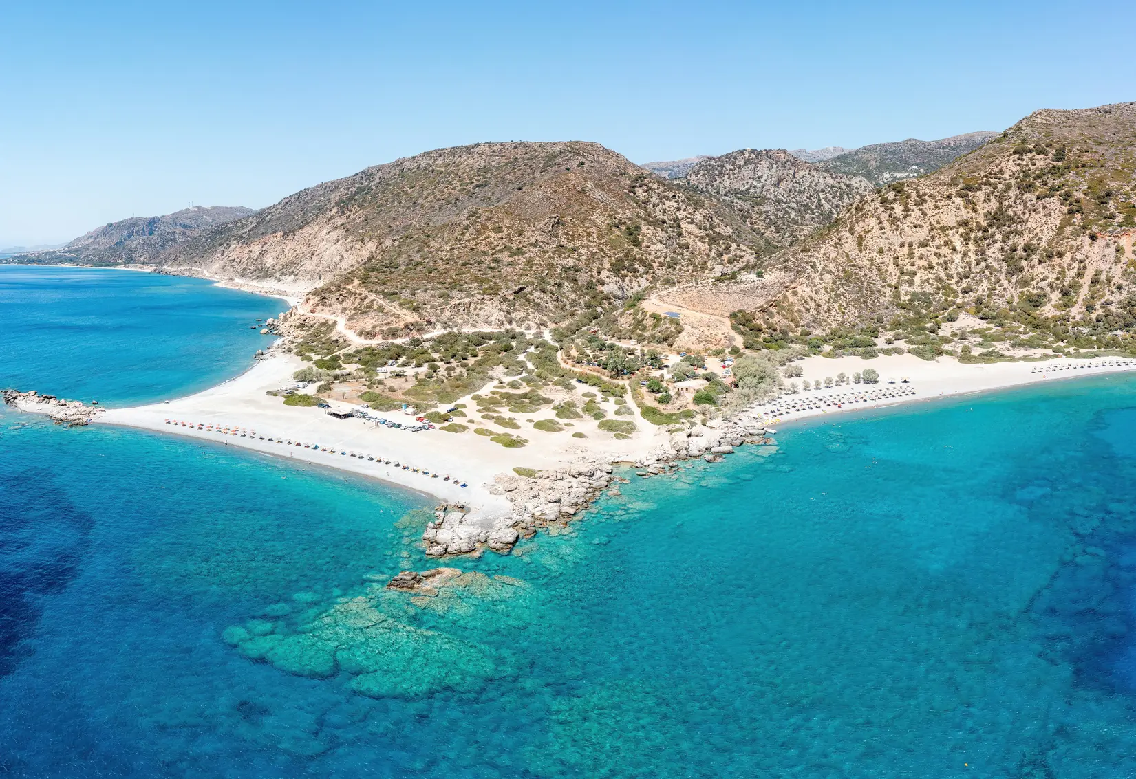 Aerial view of Gialiskari Beach in Chania, a white sand running around a headland with a row of sunbeds on each side and shallow blue water.