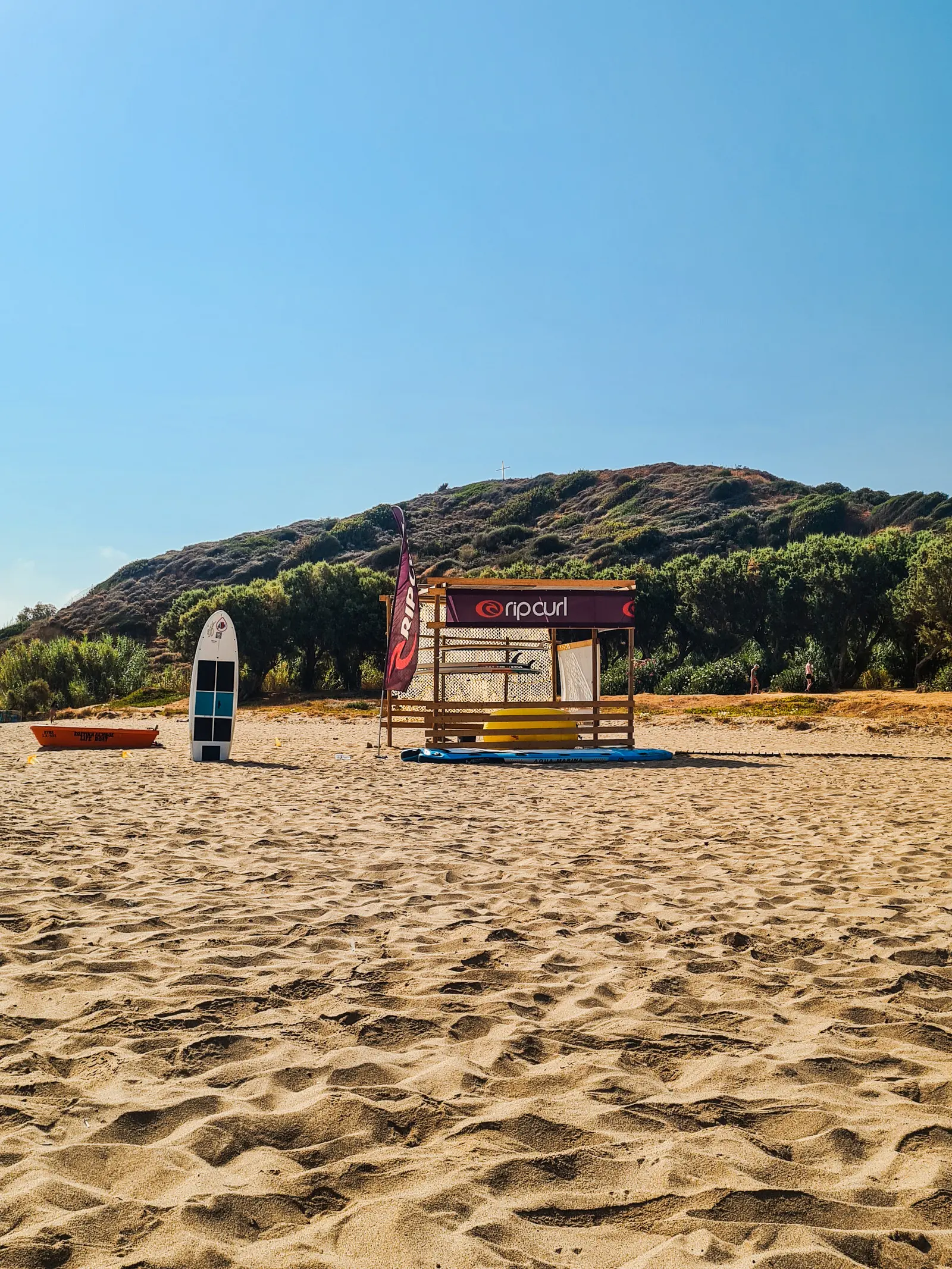 A black and white surfboard and a Rip Curl surfboard shack standing in golden sand in front of a small hill covered in greenery on Golden Beach, one of the closest beaches to Chania.