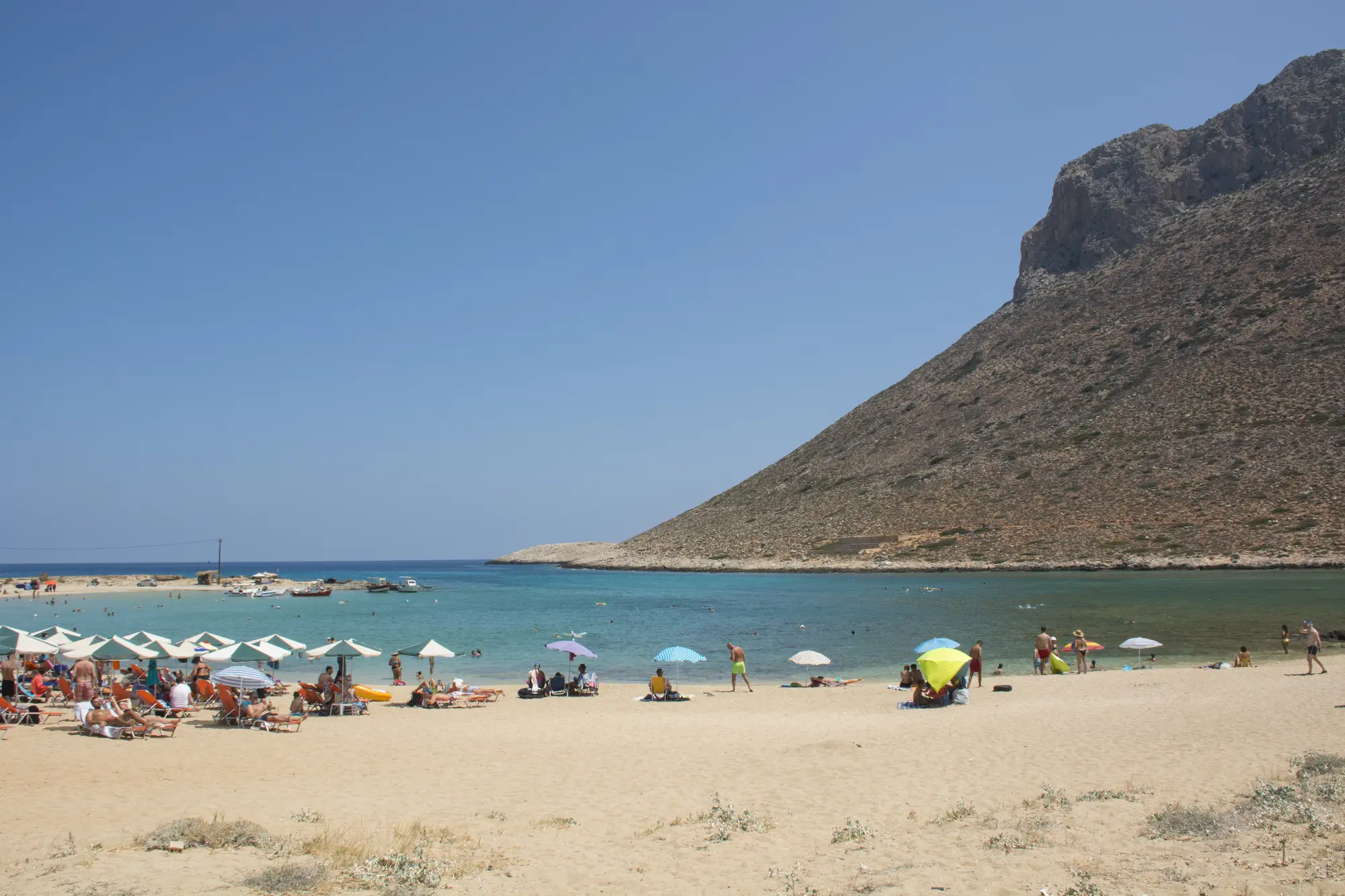 Sunbeds of a flat strip of light sand next to a tall hill and shallow blue water, Stavros Beach in Chania.