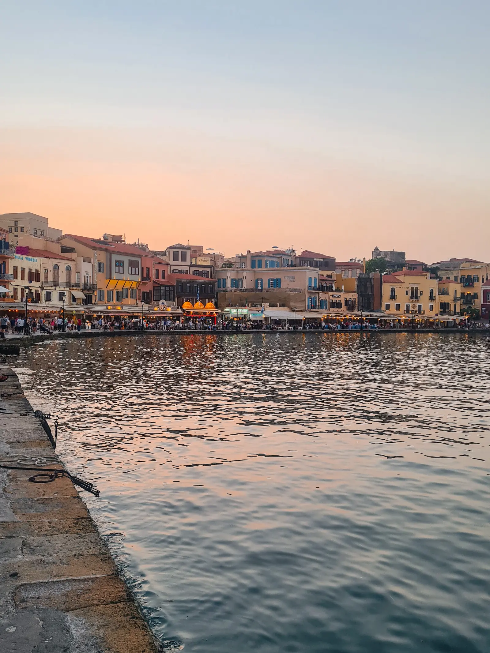 Buildings and restaurants full of people along the Venetian Harbor in Chania Old Town with an orange sunset in the background.