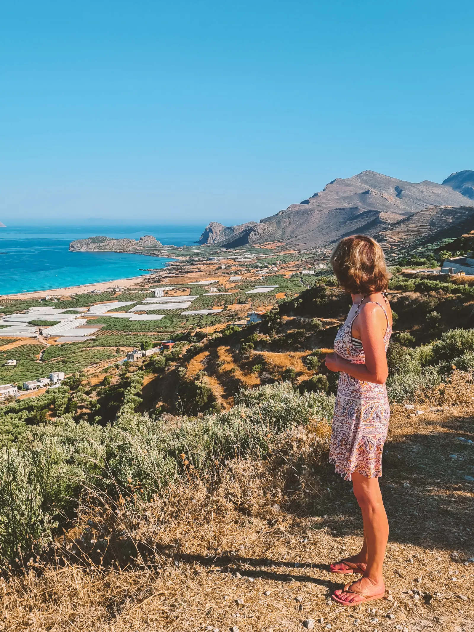 Woman wearing a short beige sundress looking out over green fields, ocean and Falassarna Beach in Crete.