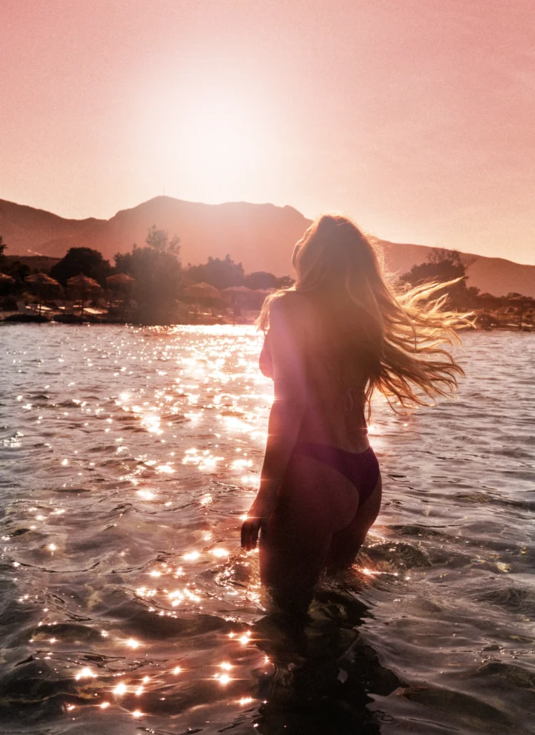 Girl with long hair, wearing a bikini, standing in the shallow water sparkling during a pink sunrise at Elafonisi Beach, the most beautiful beach in Crete.