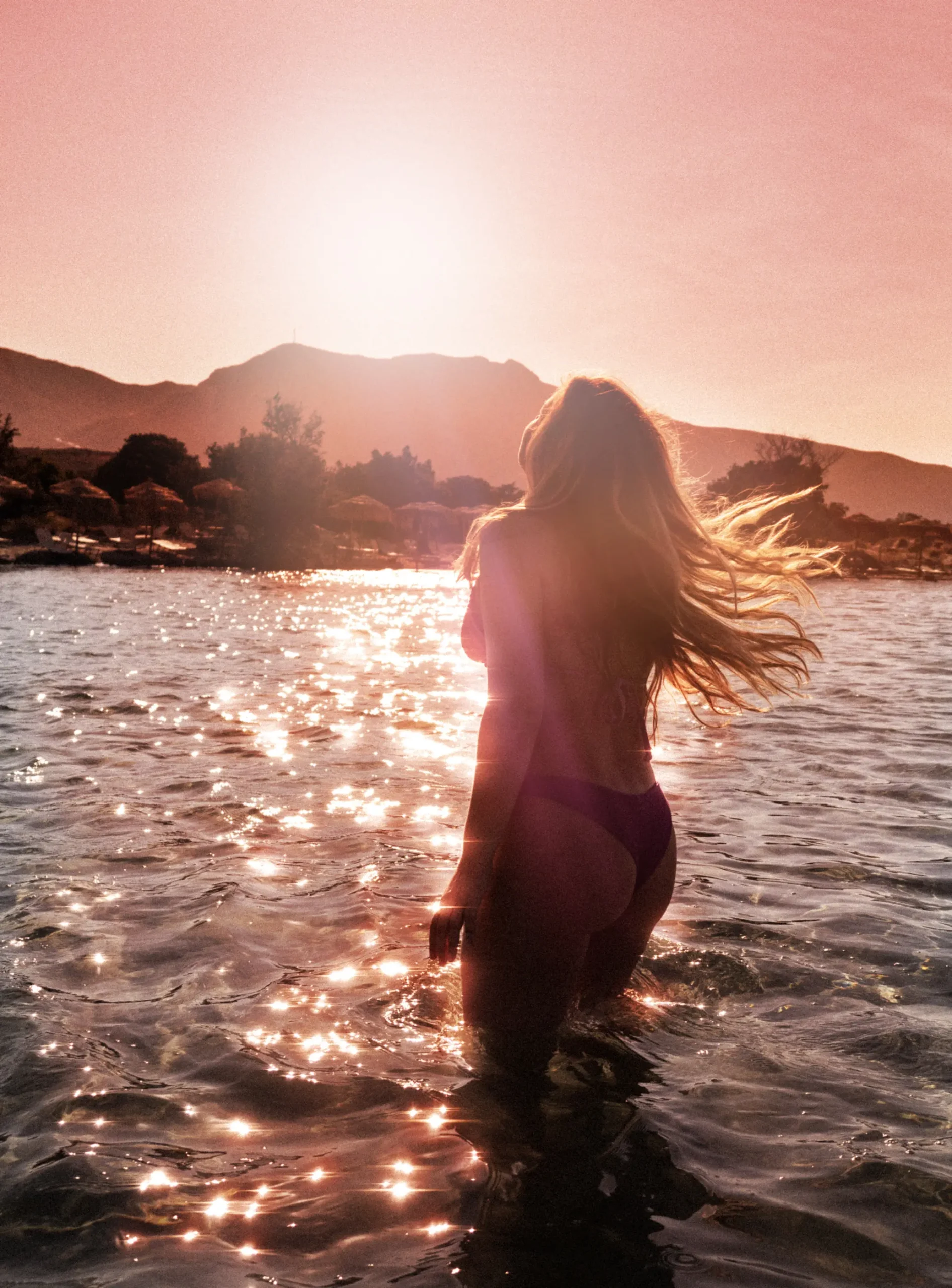 Girl with long hair, wearing a bikini, standing in the shallow water sparkling during a pink sunrise at Elafonisi Beach, the most beautiful  beach in Crete.