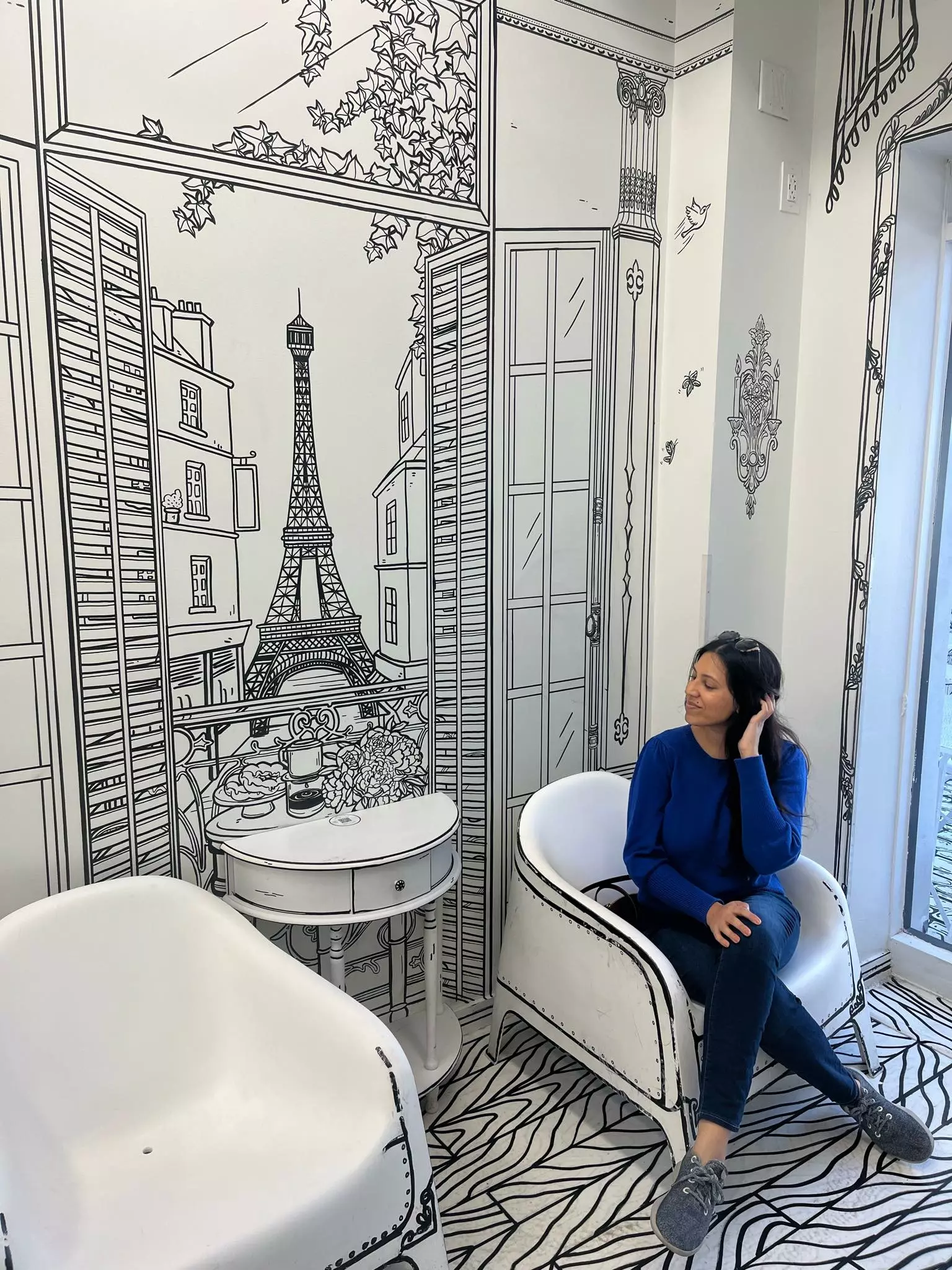 Woman wearing a blue sweater and black pants sitting in a white chair in front of a black and white Paris mural at a 2D cafe, one of the best things to do in Chicago on a weekend.