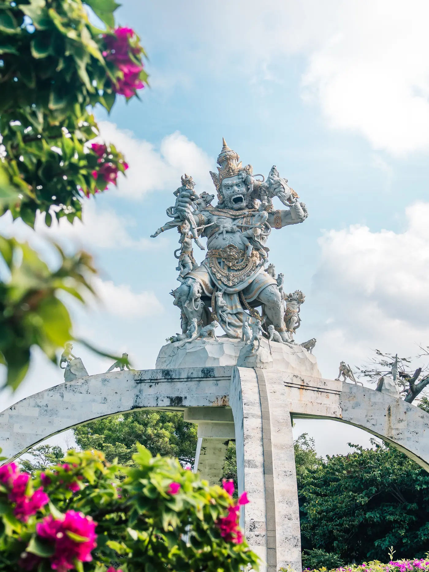 White stone Balinese god/warrior statue surrounded by greenery and purple Bougainvillea at Uluwatu Temple in Bali.