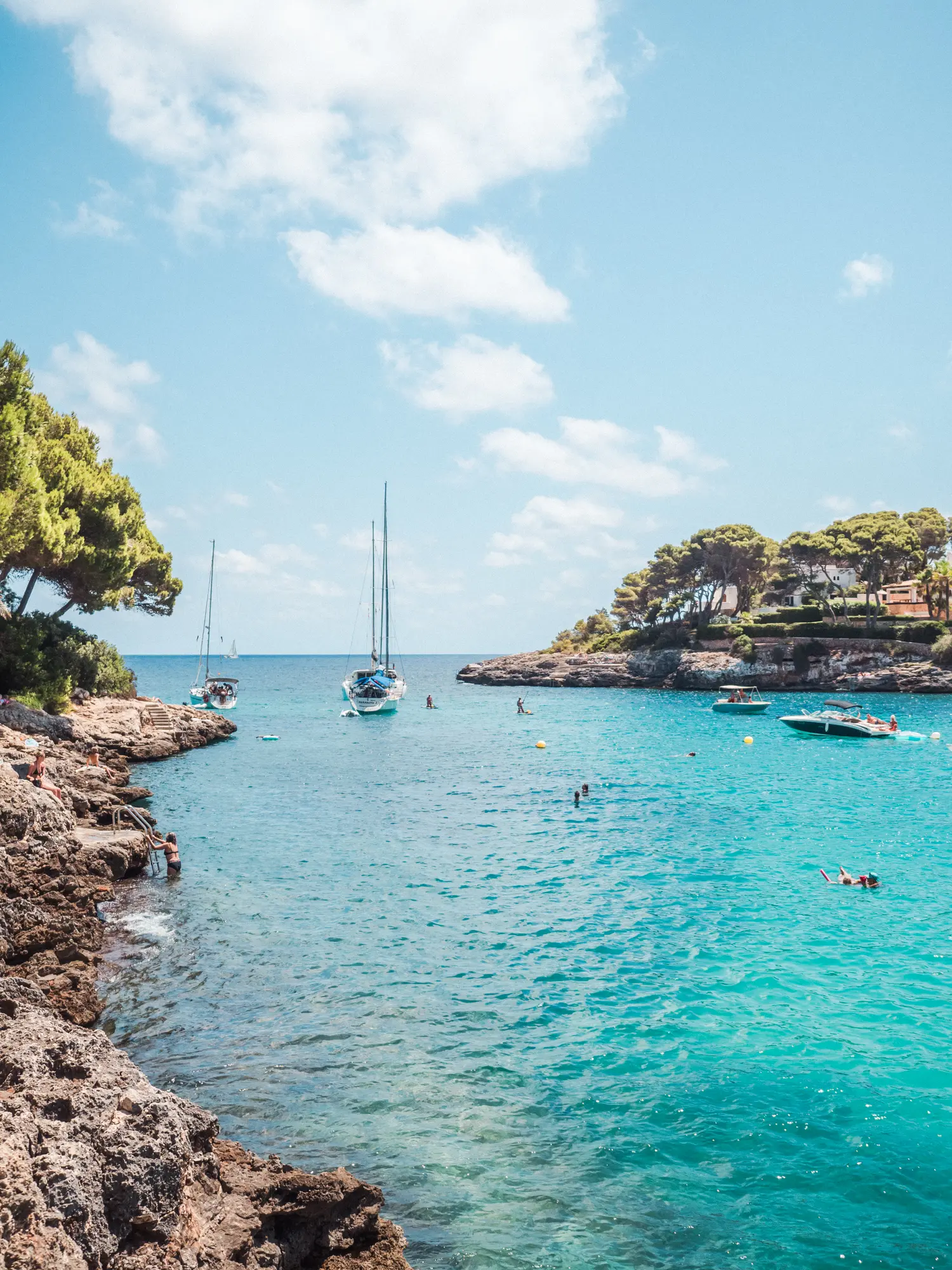 Two sailboats and two smaller boats with people swimming around in the turquoise ocean of Cala Gran with rocks on each side, one of the best beaches in Mallorca.
