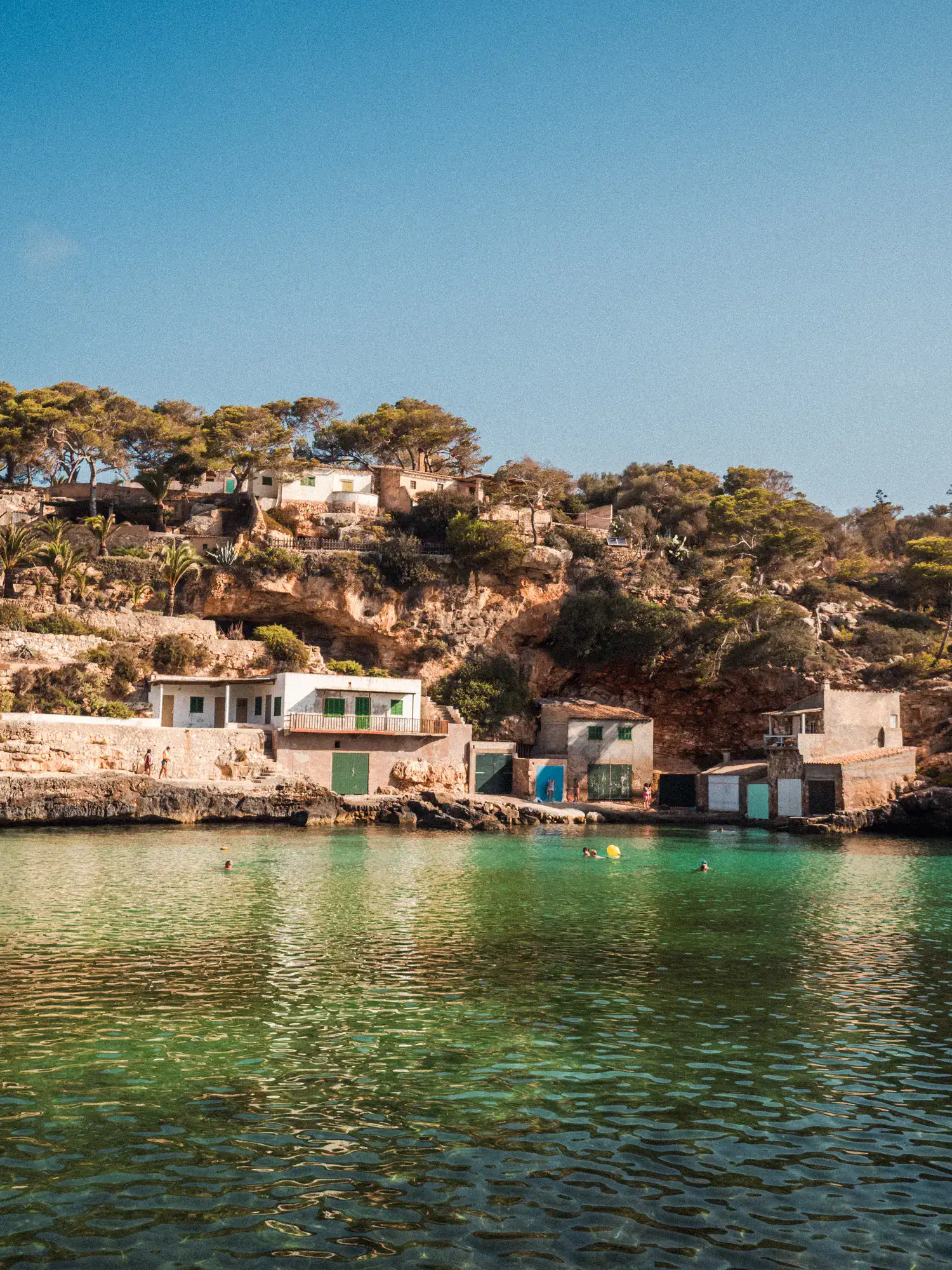Rustic white and stone boathouses with green shutters nestled along the rocky cliffs of Cala Llombards in Mallorca, Spain, with crystal-clear turquoise water and swimmers enjoying the calm cove under a clear blue sky.