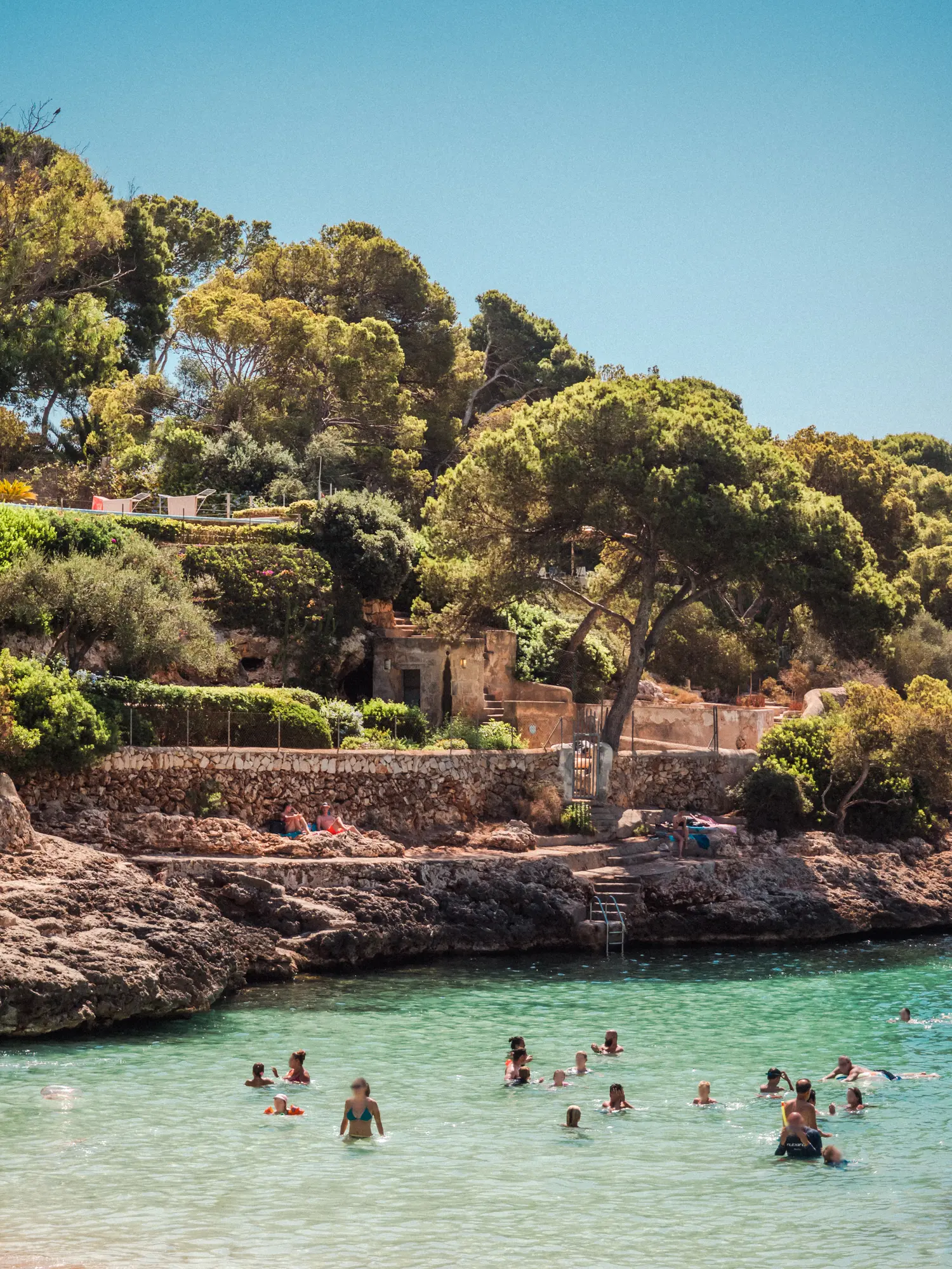 People in shallow green water with rocks and greenery in the background, Cala D'Or Mallorca.