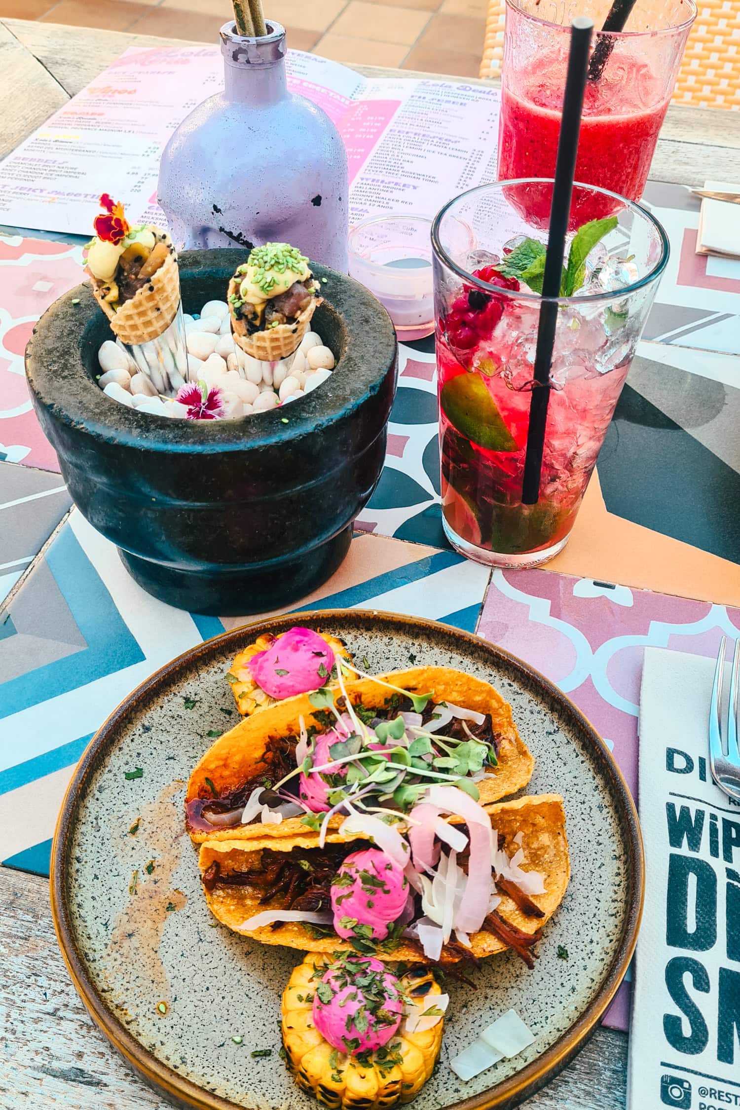 Green and brown plate with pulled pork tacos with pink sauce, strawberry mojito and tuna tartar on a colorful table at Diferent Restaurant in Cala d'Or Mallorca.