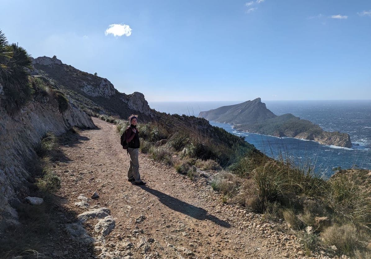 Woman wearing dark hiking gear and a backpack hiking on a dirt overlooking the ocean in Serra de Tramuntana Mountains in Mallorca.