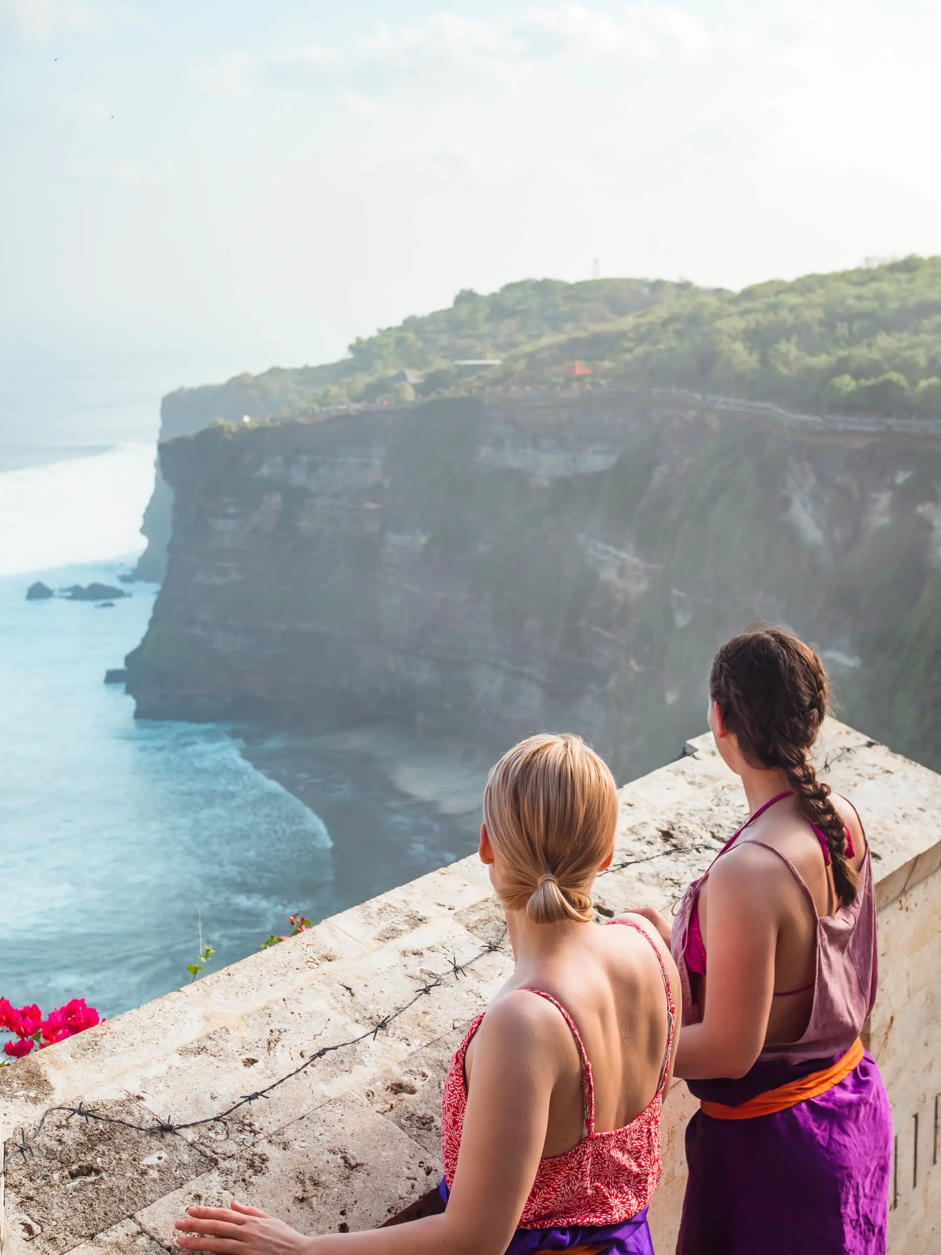One blonde snd one brunette girl wearing purple sarongs looking over the wall at the ocean and surrounding cliffs at Uluwatu Temple.