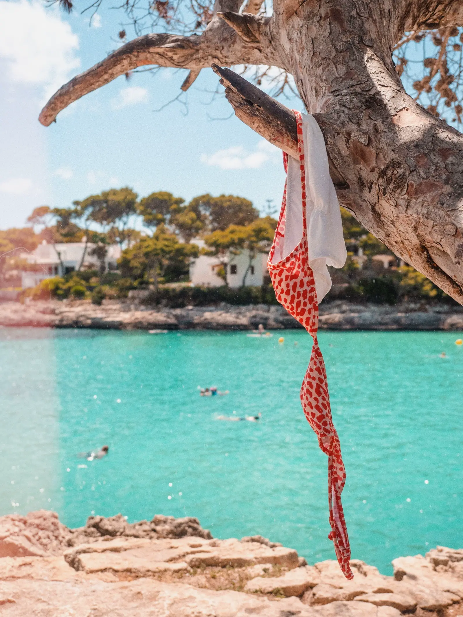 Red and white swimwear hanging on a tree in front of turquoise water of Cala Gran, one of the best beaches in Cala d'Or Mallorca.