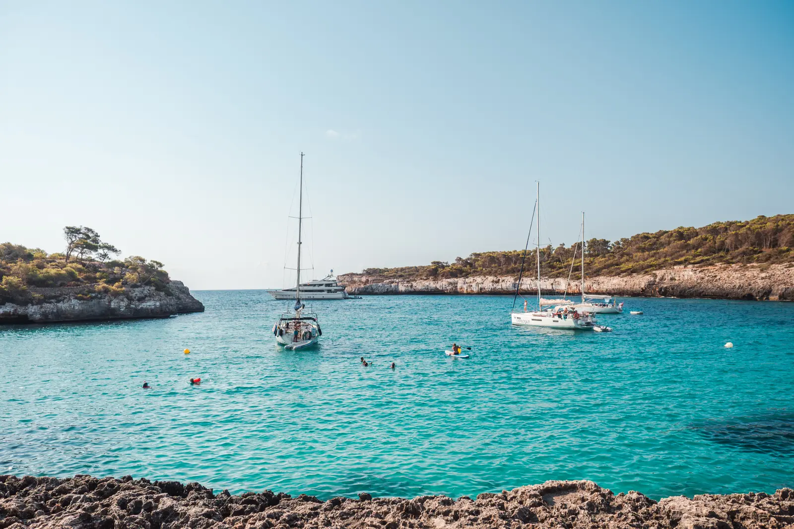 Three white sailboats and a yacht with people swimming around in the turquoise ocean of Cala Mondrago Mallorca.