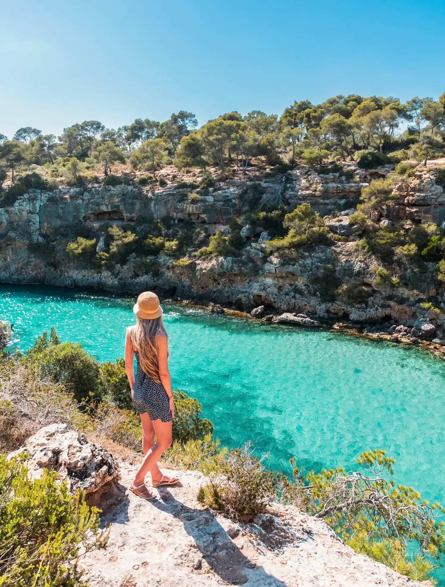 Woman with long hair, wearing a black and white pattern dress and beige hat, standing in a cliff looking down on the turquoise water of Cala Pi in Mallorca.